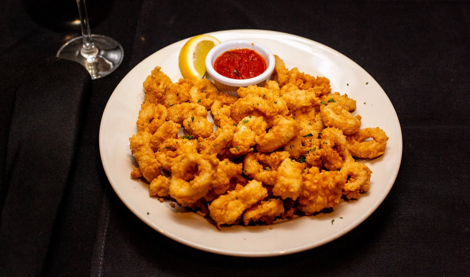 Plate of fried calamari with a lemon wedge and a small bowl of marinara sauce on a white plate, black tablecloth background.