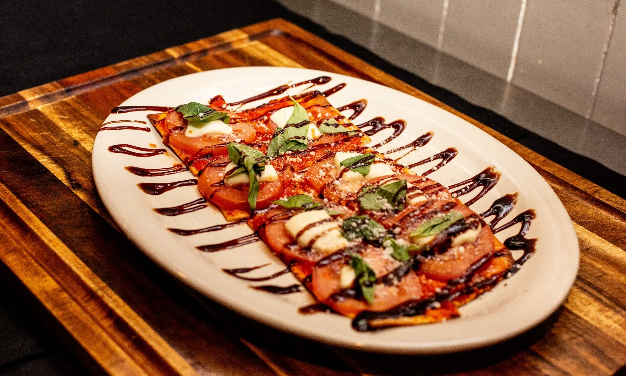 A flat bread pizza with tomato slices, basil leaves, mozzarella cheese, and drizzled balsamic glaze, served on a white oval plate on a wooden board.
