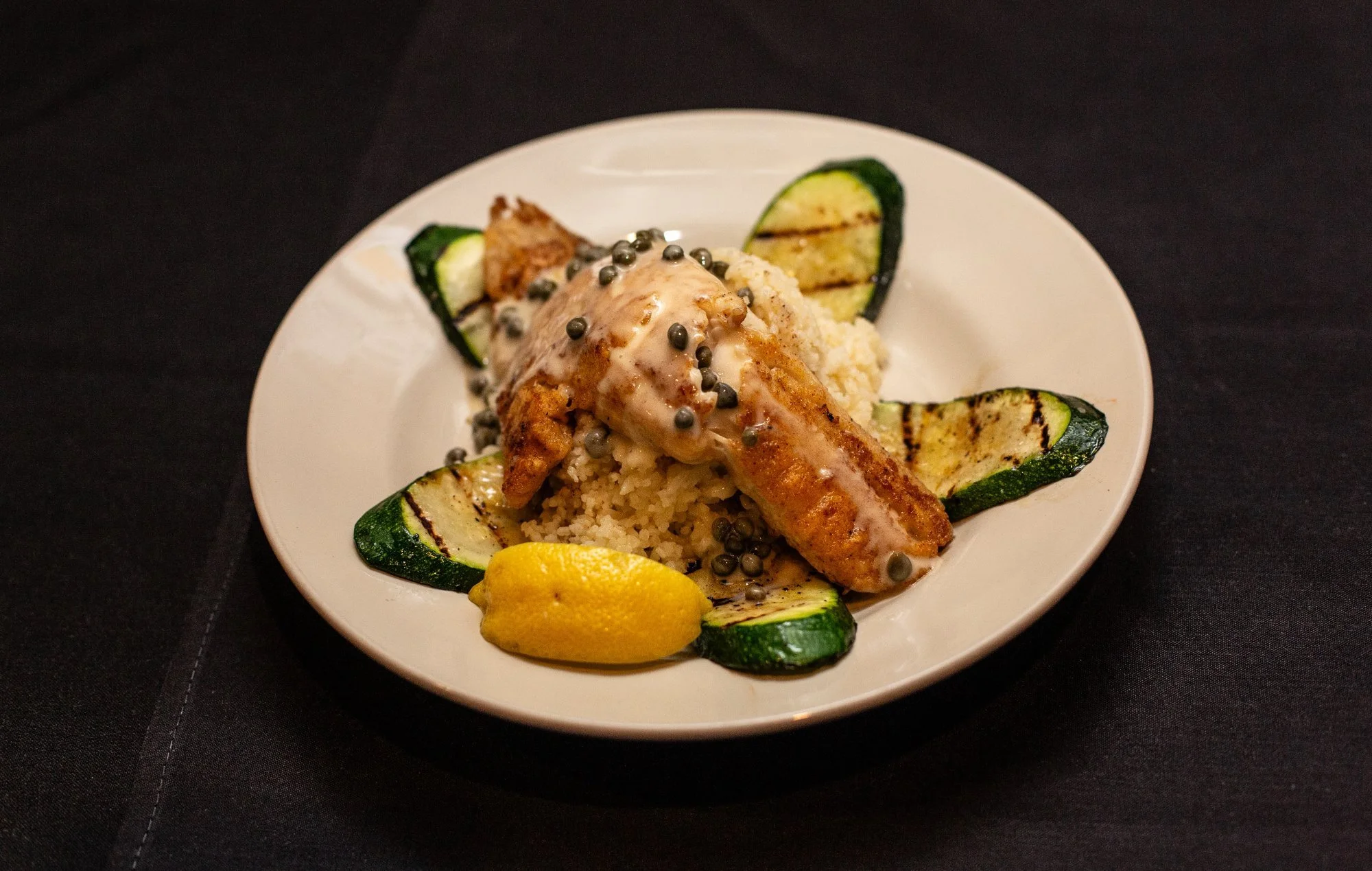 Plate with grilled zucchini slices, lemon wedge, and a serving of seasoned chicken topped with sauce and black pepper pearls, on a black table.