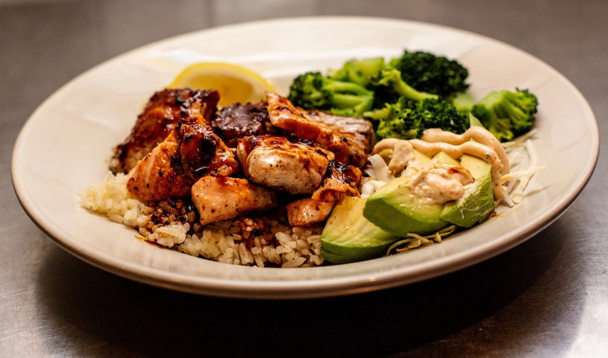 Plate of grilled chicken, rice, broccoli, avocado, and a lemon wedge.