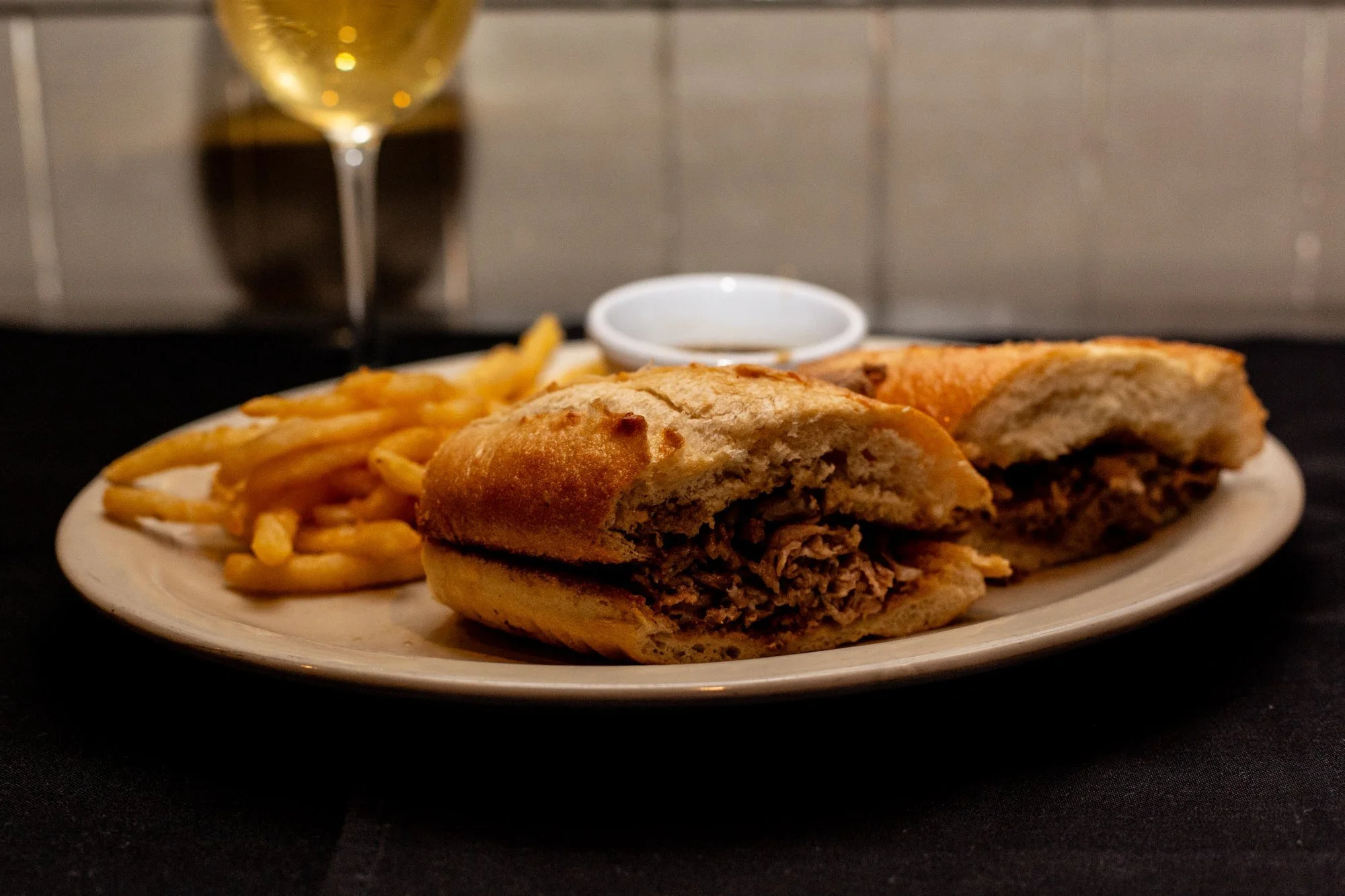 A plate of French fries and a beef sandwich with shredded meat, served with a side of dipping sauce and a glass of white wine in the background.