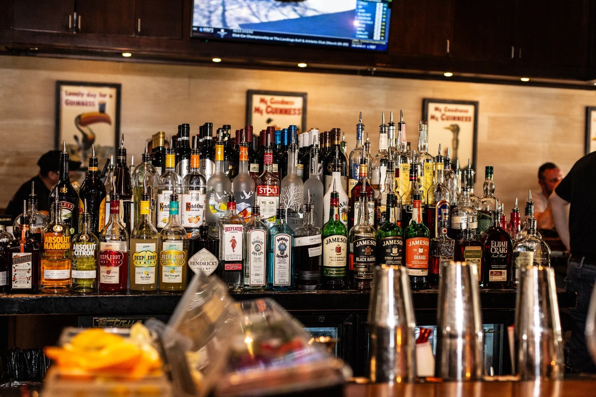 Bar with a variety of liquor bottles, including Jameson Irish Whiskey, Hendrick's Gin, and Absolut Vodka, arranged behind the counter in a restaurant or bar setting, with patrons and bartenders in the background.