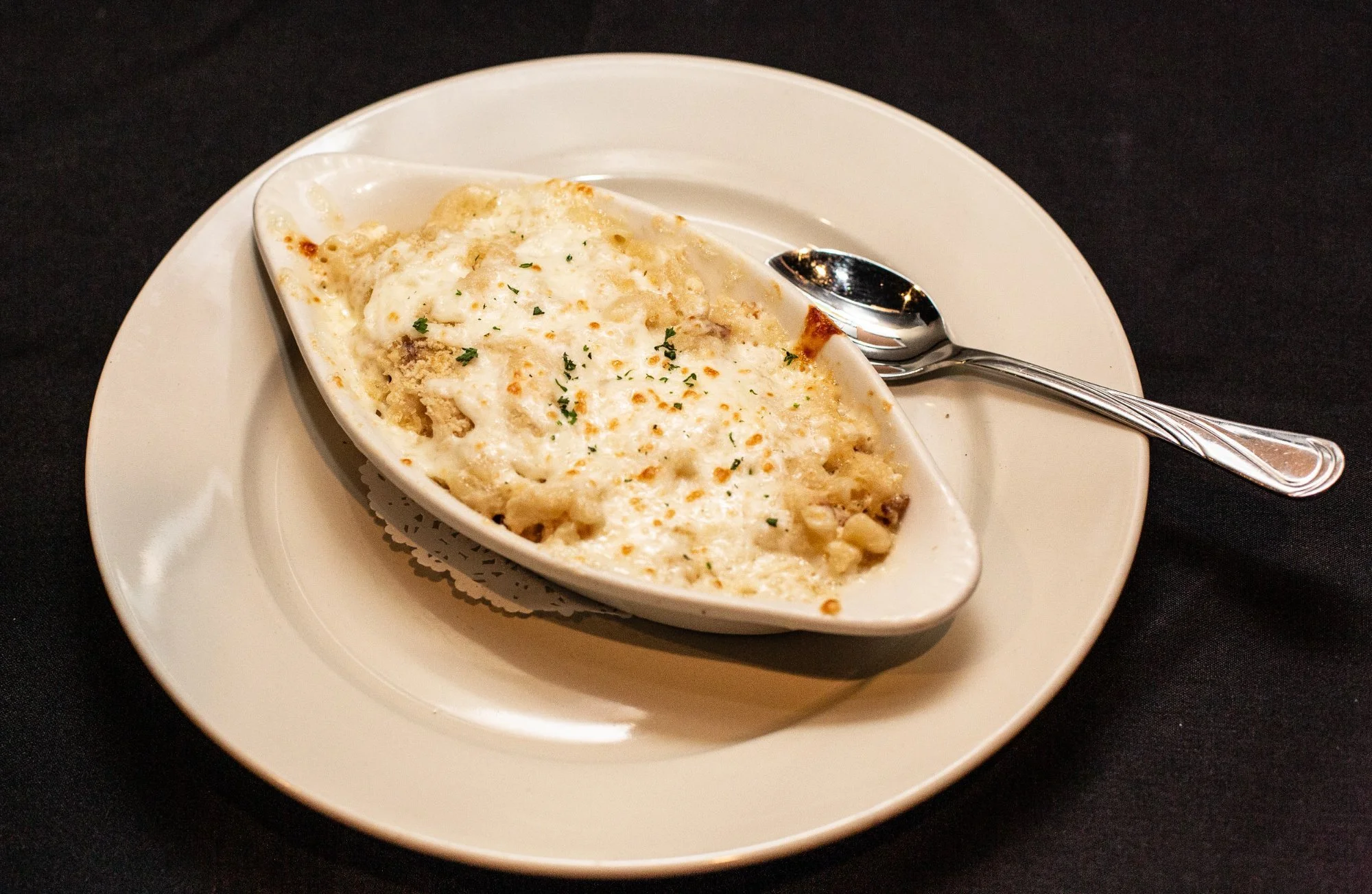 Baked macaroni and cheese in a white oval dish on a white plate with a spoon, set on a black table.