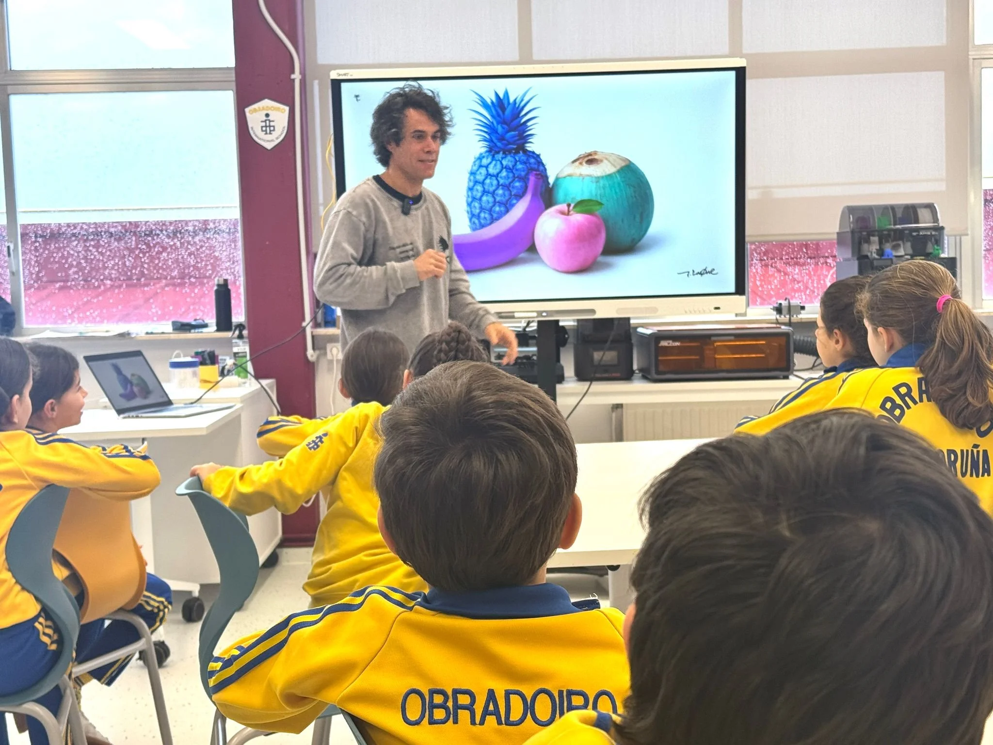 Javi Luque como profesor enseña a un grupo de niños sobre storytelling creativo sobre colores, mostrando en una pantalla imágenes de fruta tropical como piña, plátano, manzana y coco.