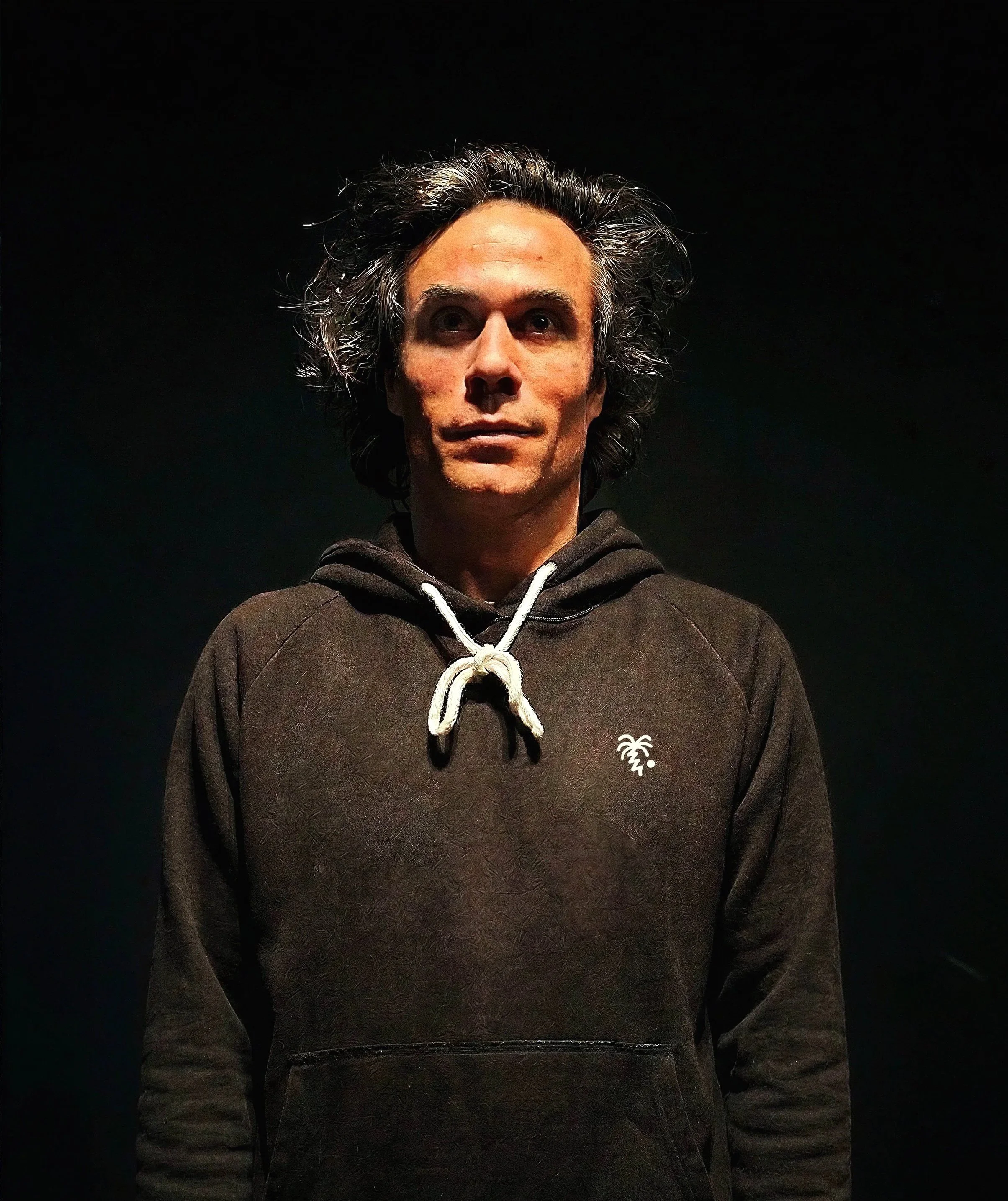 Javi Luque director creativo, de estrategia de marca y artista vistiendo sudadera negra con capucha y dibujo de una palmera, en un fondo negro.