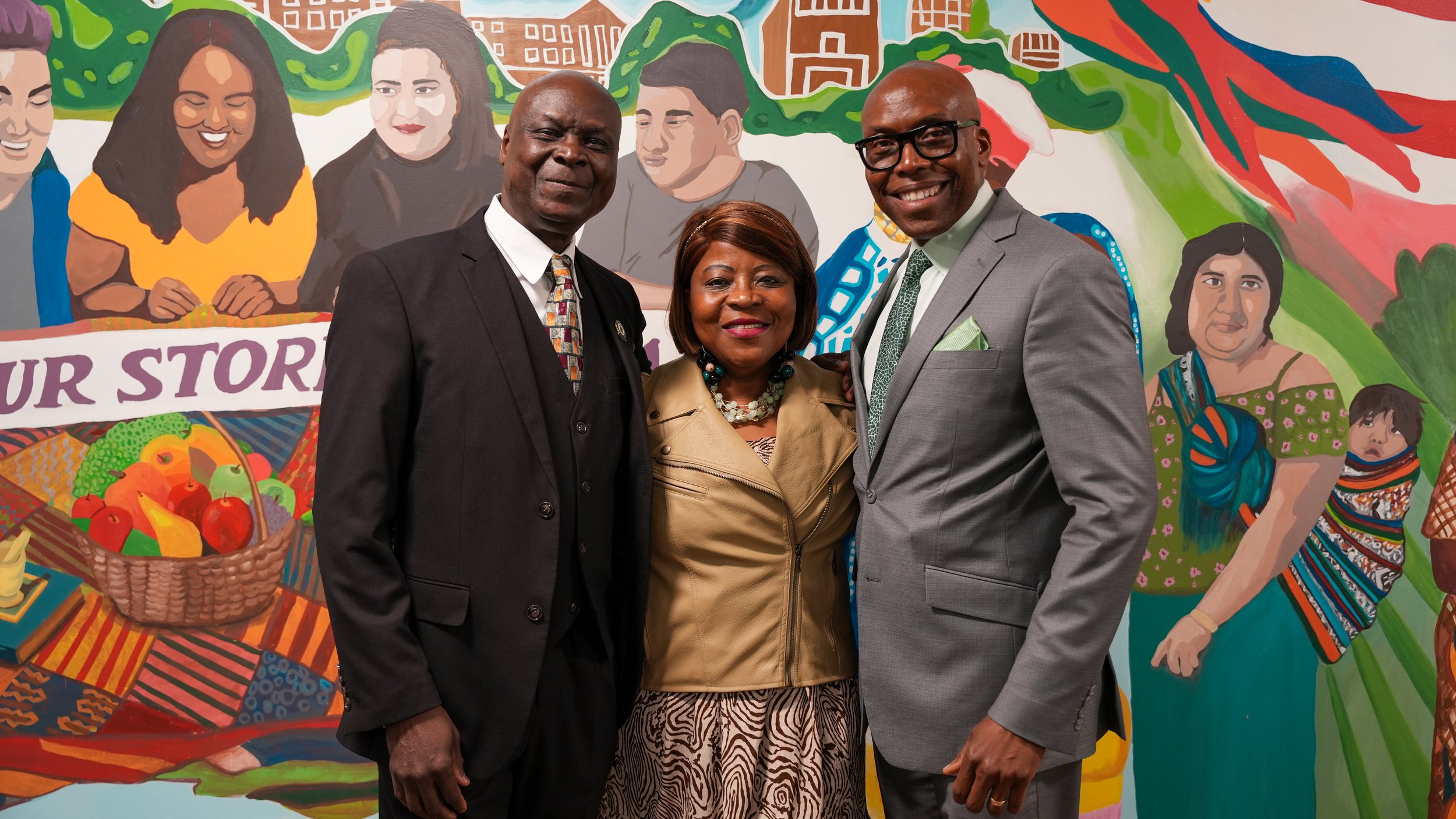 Three adults standing together in front of a colorful mural featuring diverse faces, fruits, and greenery, smiling for the photo.