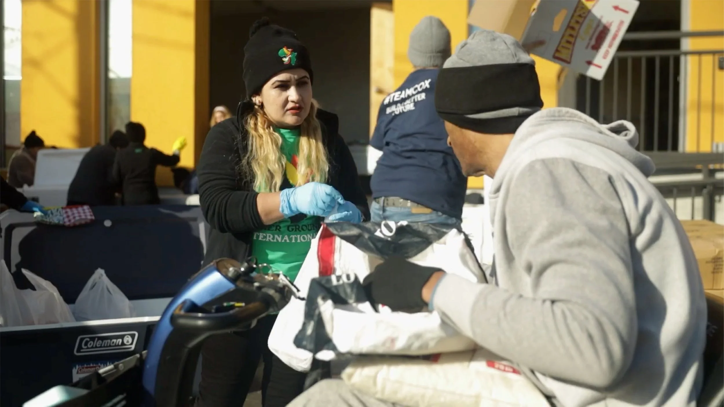 A woman with long blonde hair, wearing a black beanie with a colorful logo and a green shirt, is handing out items to a man in a gray hoodie and black beanie at an outdoor event. There are tables with bags and other people in the background.