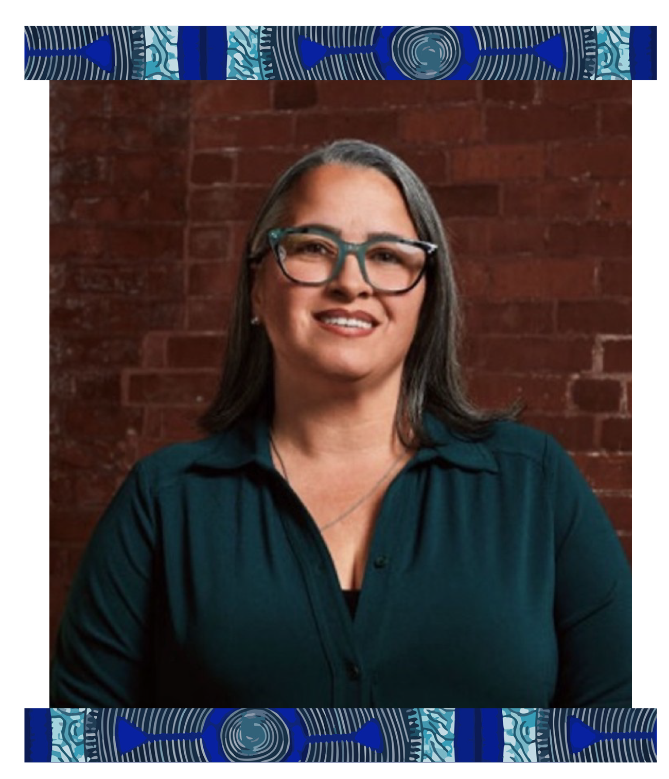 A woman with glasses, dark hair with gray streaks, smiling, wearing a dark teal blouse, posed against a brick wall.