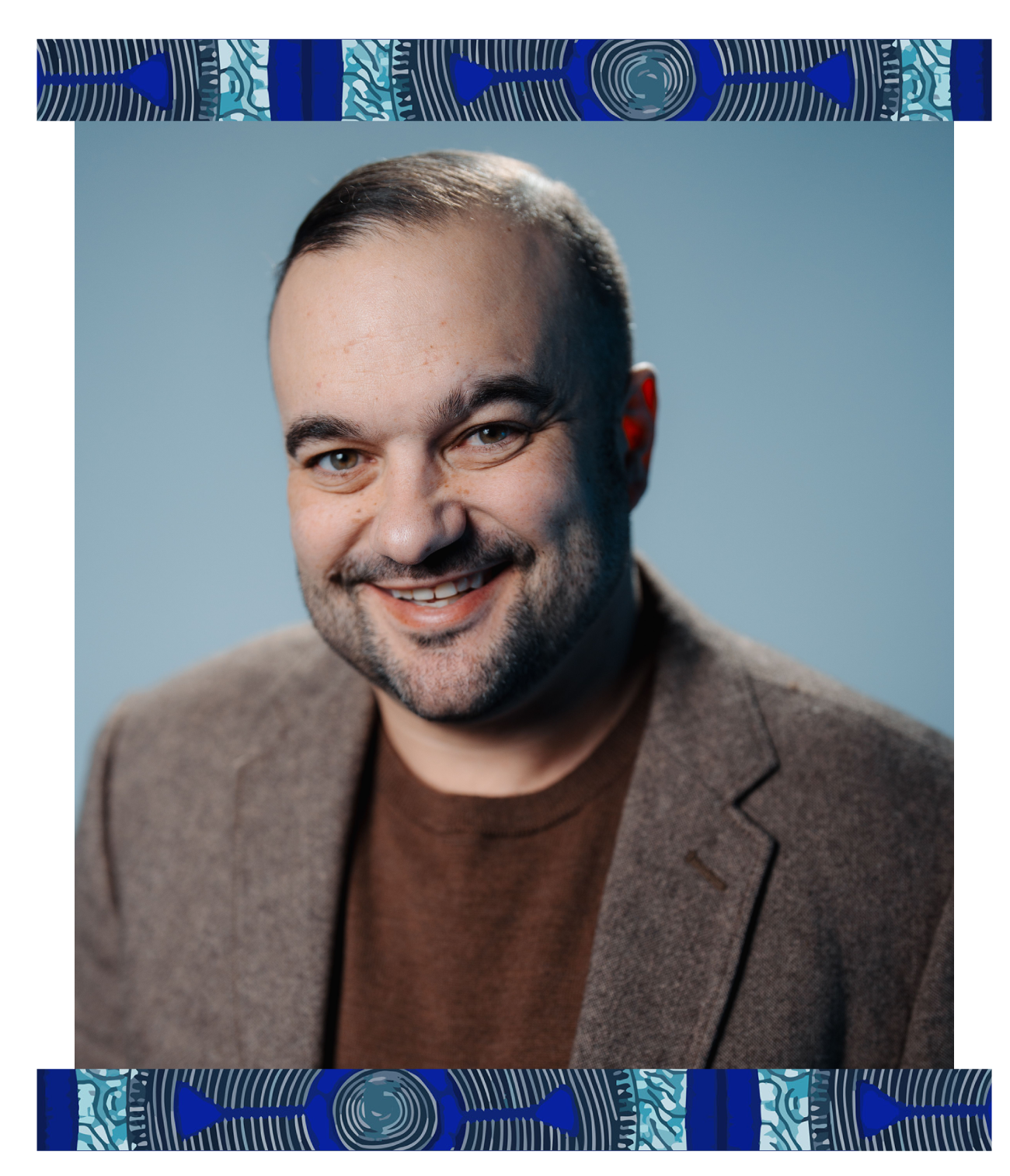 Portrait of a smiling man with a beard and short dark hair, wearing a brown blazer and a brown shirt, against a plain blue background.