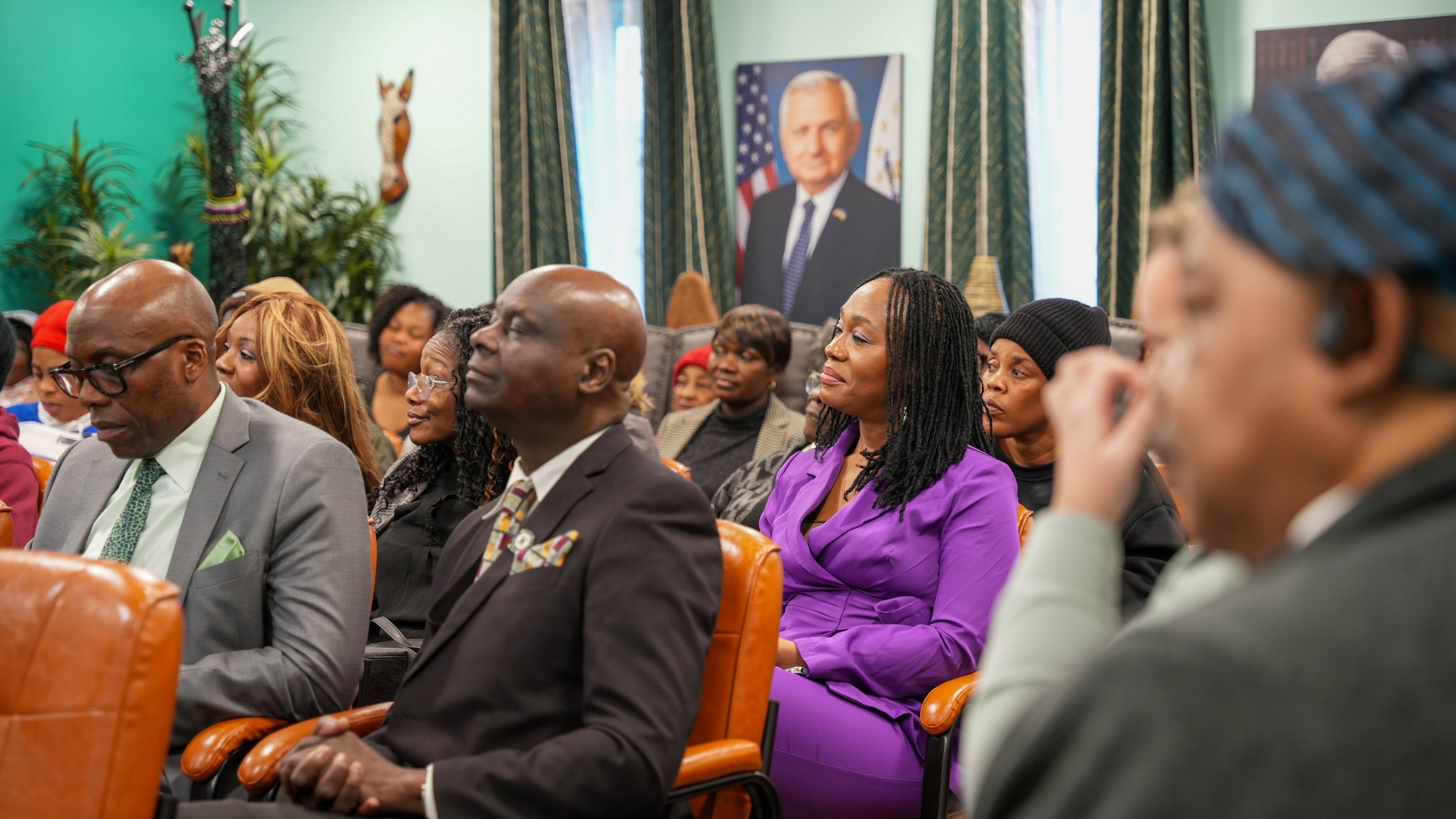A group of diverse people attending a formal event or meeting, seated in rows in a room decorated with plants and patriotic portraits, including one of a man with a flag background.