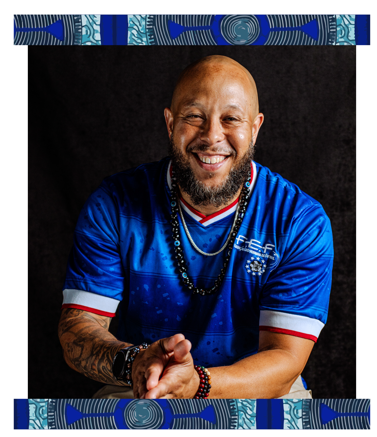 A smiling man with a bald head and beard, wearing a blue sports jersey with white and red accents, accessorized with necklaces and bracelets, posing against a black background.