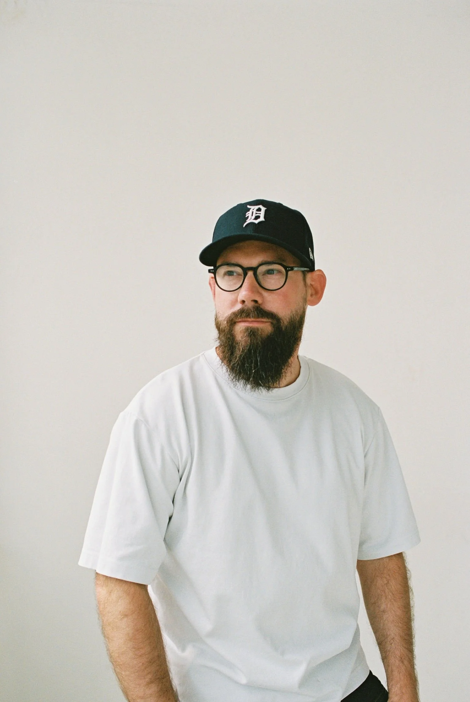A man with glasses and a beard wearing a white t-shirt and a black baseball cap with a white logo, standing in front of a plain light-colored wall.