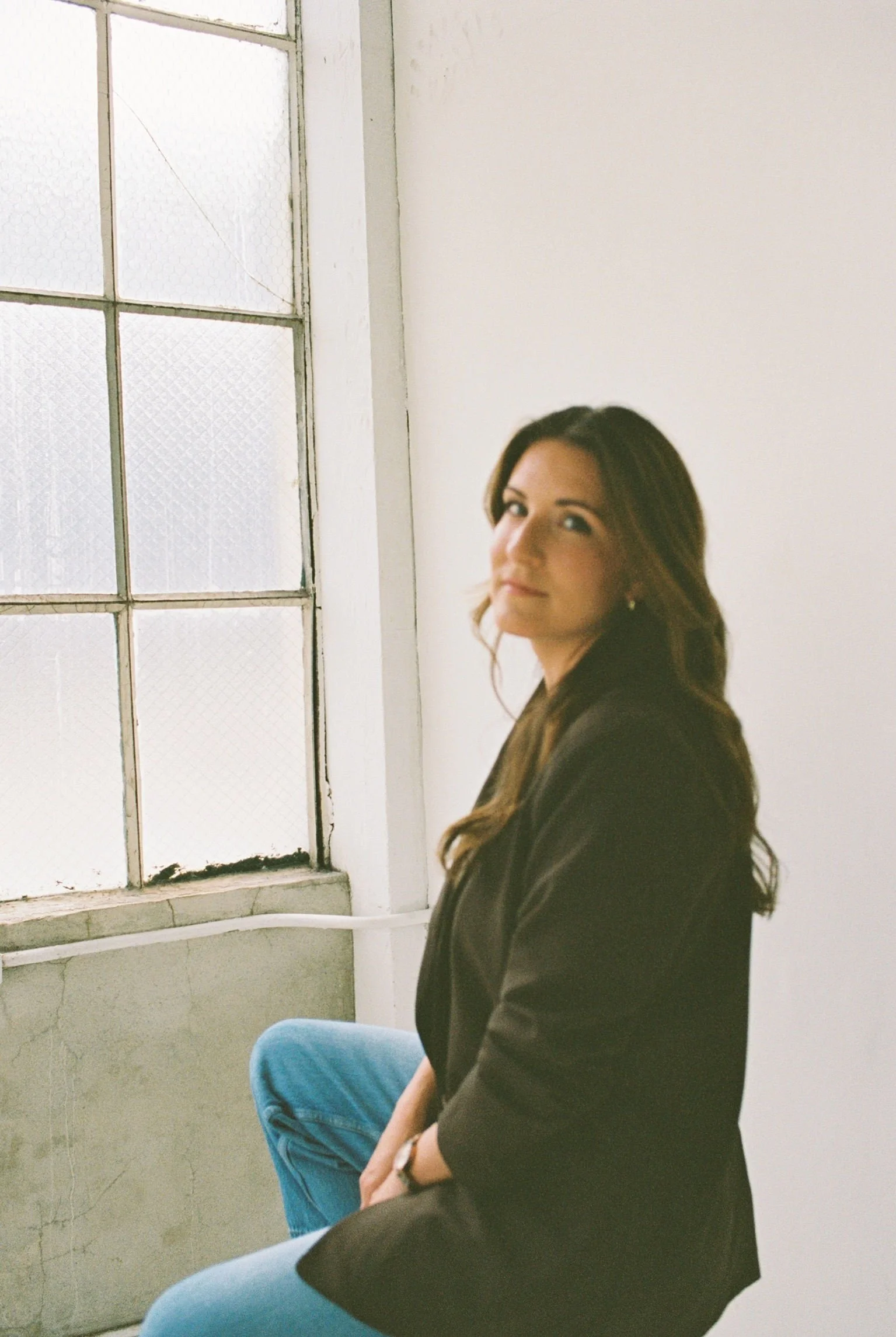 A woman with long brown hair and a black jacket sitting in front of a large window with frosted glass panes. The window has a white frame and a ledge, with some dirt or damage visible at the bottom. The woman is sitting next to a person in a blue shirt.