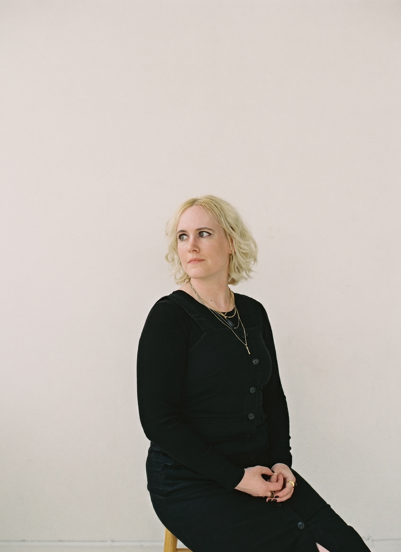 A woman with blonde curly hair, wearing a black button-up shirt with layered necklaces, sitting on a stool against a plain white wall.