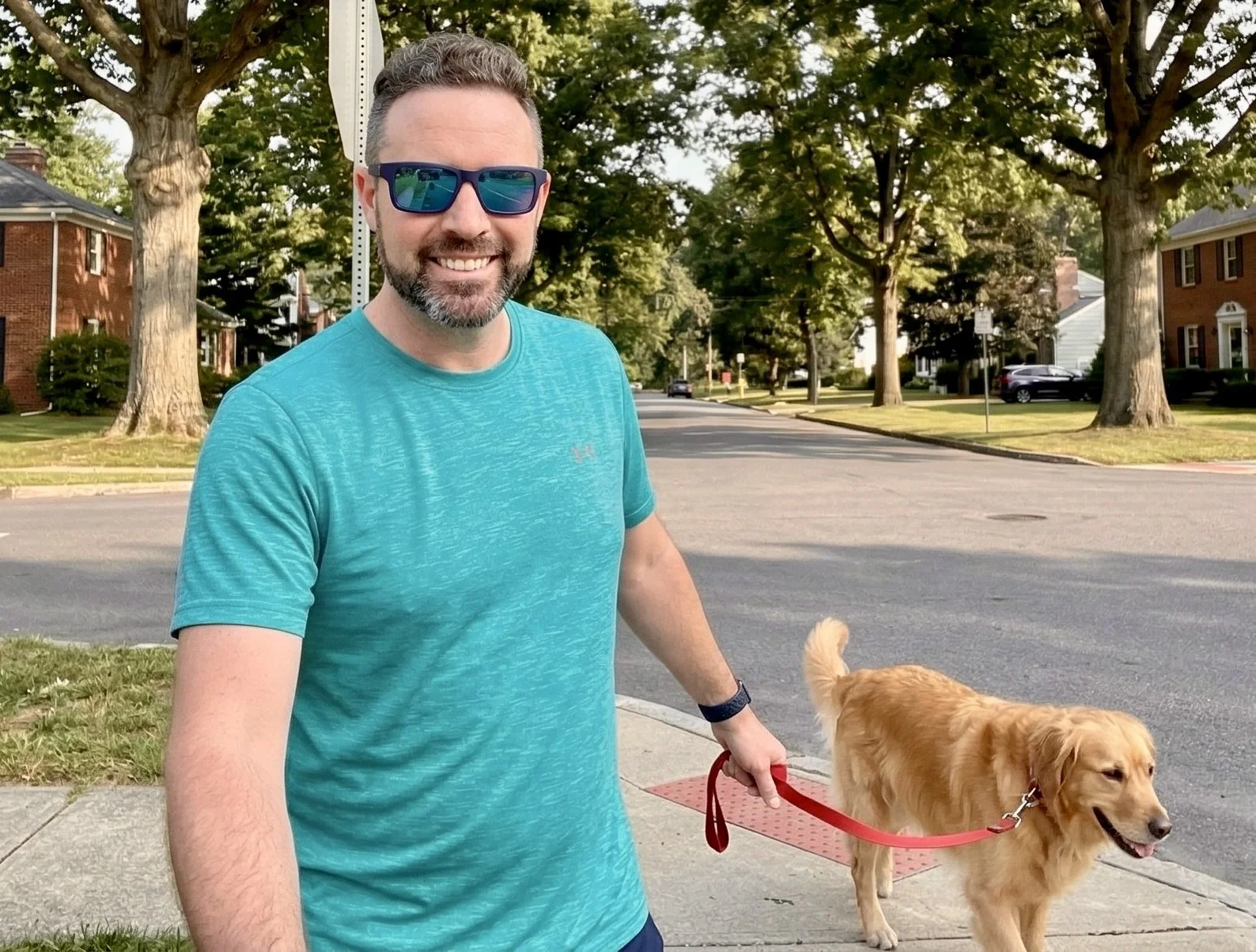 A man with dark hair and a beard, wearing sunglasses and a turquoise T-shirt, smiling while walking a golden retriever on a red leash in a residential neighborhood with trees and houses.