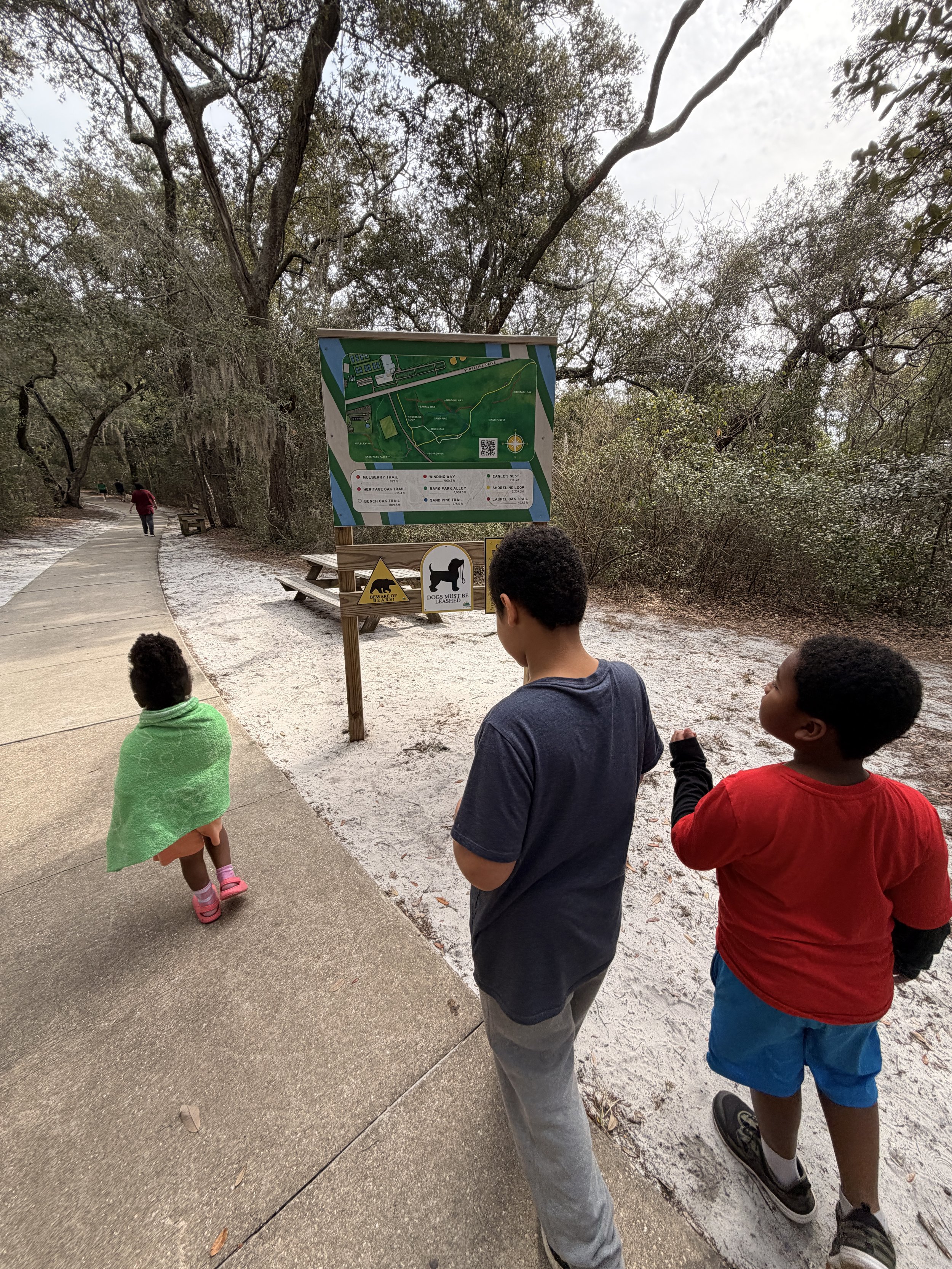 Three children on a nature trail looking at a park map and signs at a wooded park area.