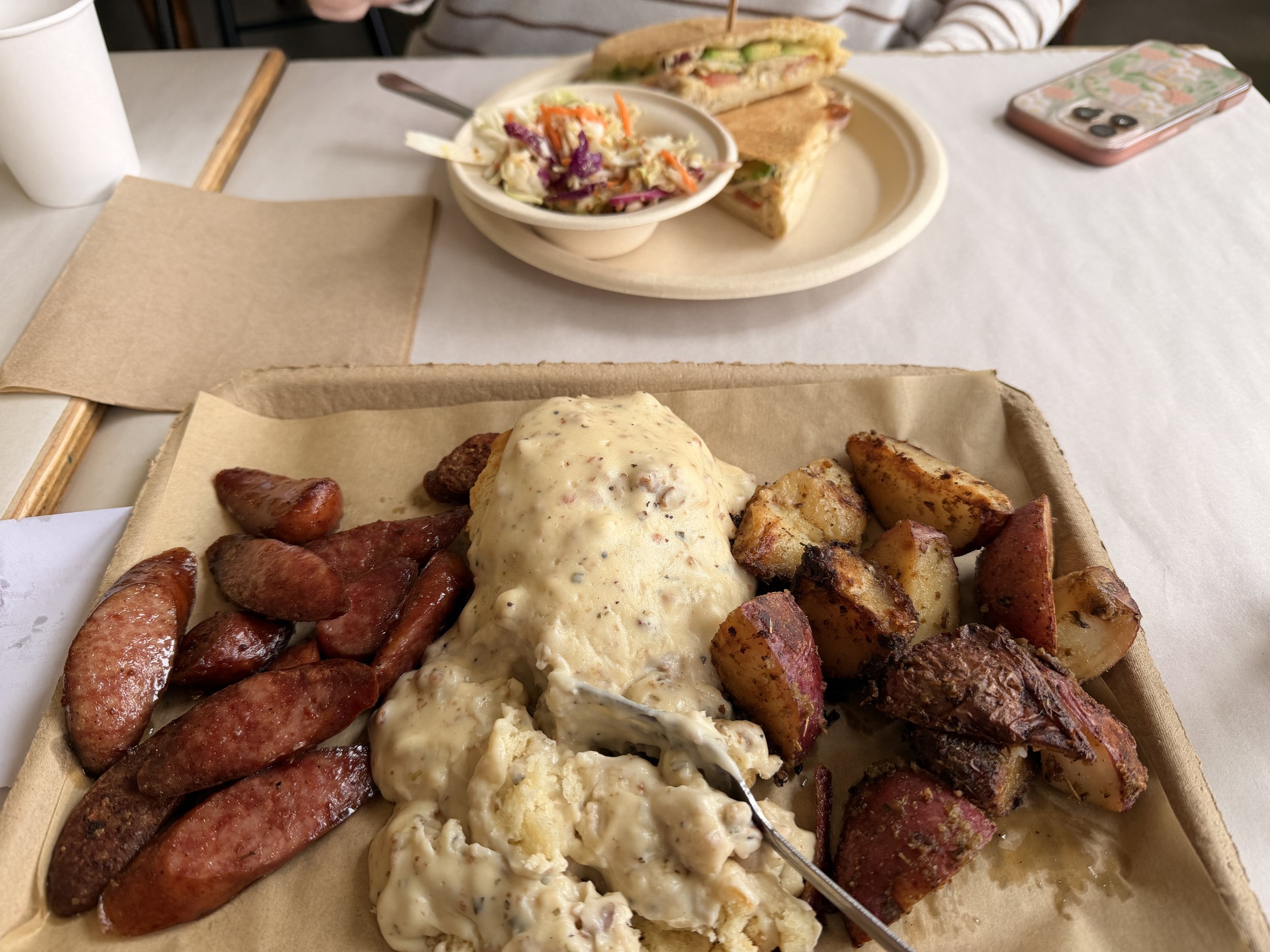 Plate of breakfast food including scrambled eggs, sausage links, roasted potatoes, and sausage gravy, with a fork resting on the plate. In the background, there is a sandwich, a bowl of coleslaw, a cup, and a smartphone on a table.