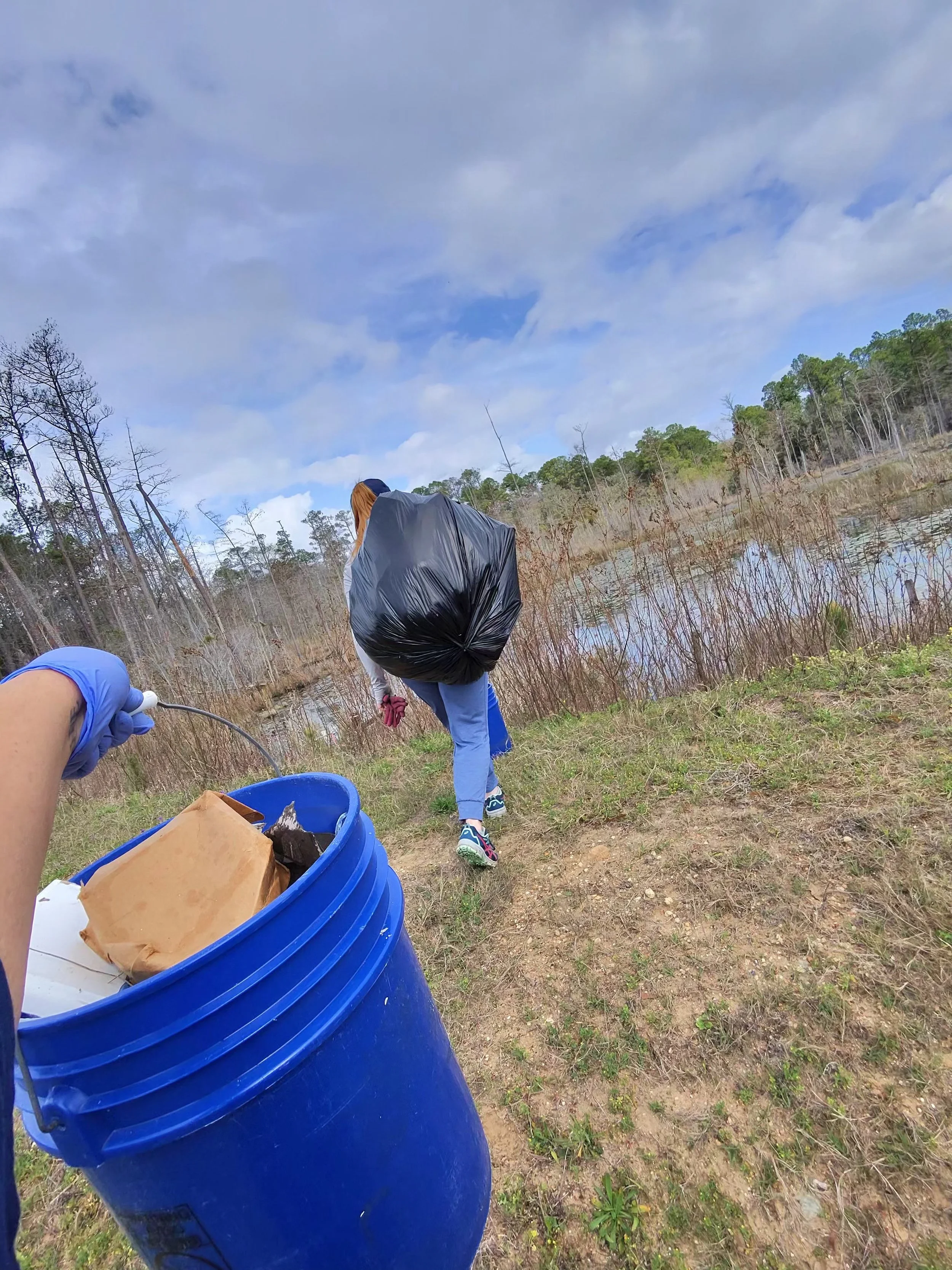 Someone in blue gloves holding a blue bucket with trash, following a person with a large black trash bag near a wetland area on a cloudy day.