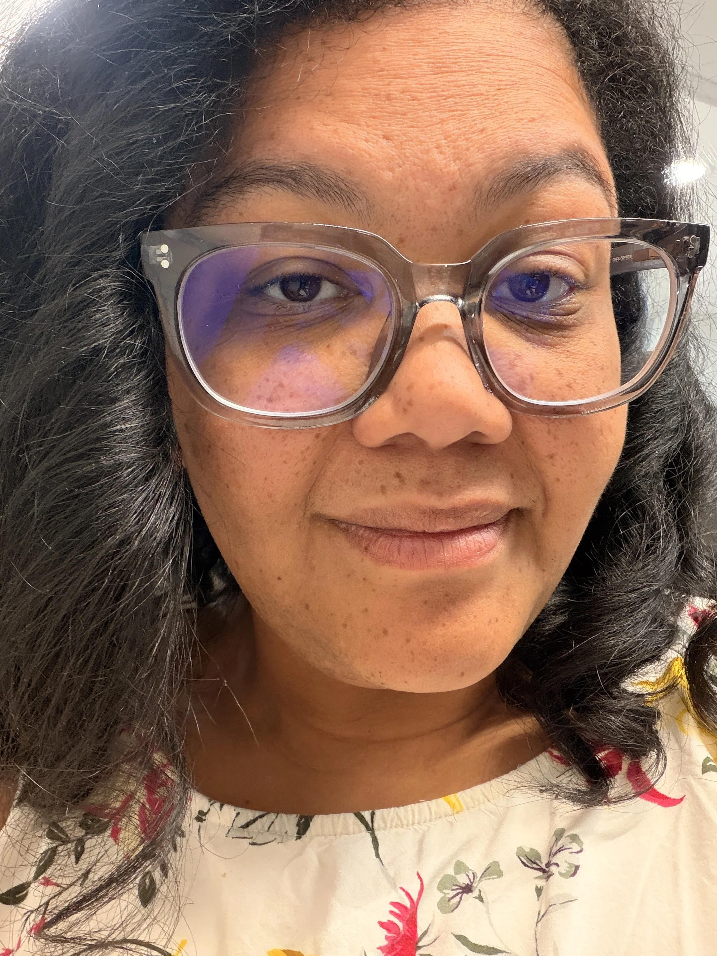 A woman with curly black hair wearing glasses and a floral top, smiling at the camera. This is Tyler L. Davis.