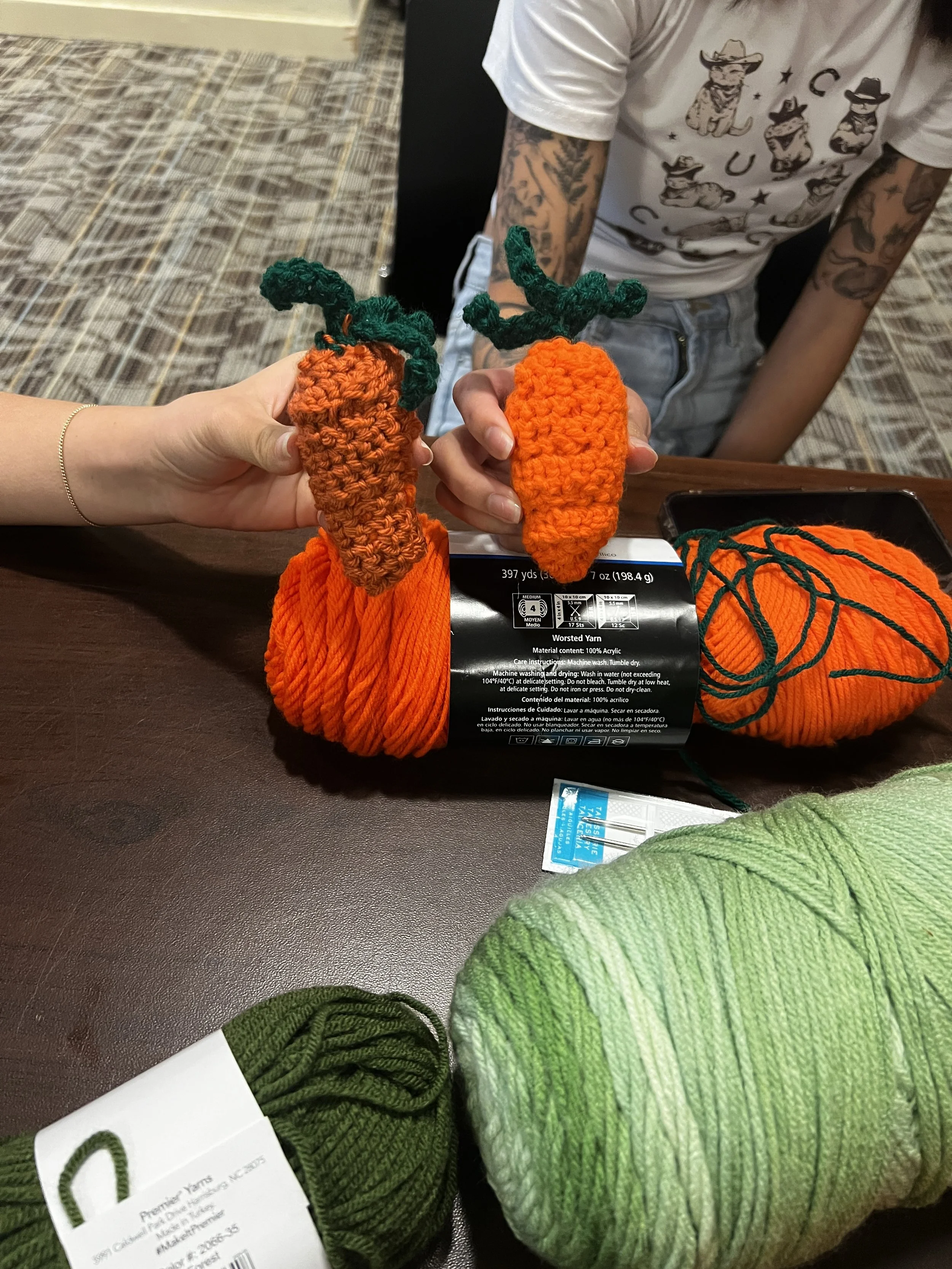 Two crocheted carrots with green stems, one held by a person with lighter skin and the other by a person with darker skin, placed on a table with yarn and knitting supplies.