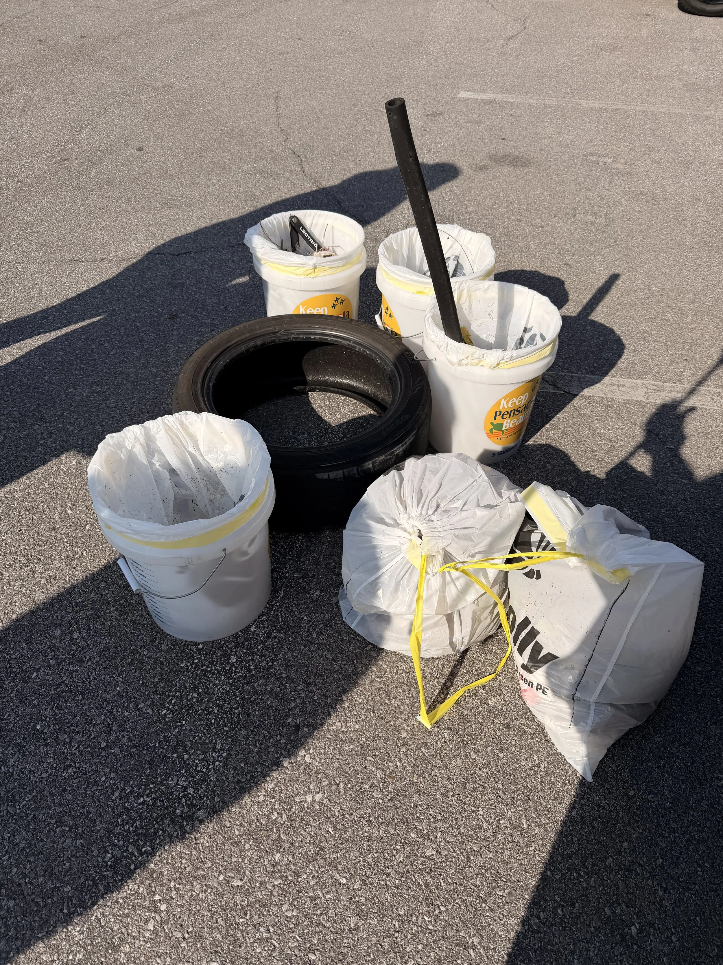 Outdoor scene with discarded trash including a black tire, five white trash bags, and a black pole, all on asphalt ground, with shadows cast across the area.