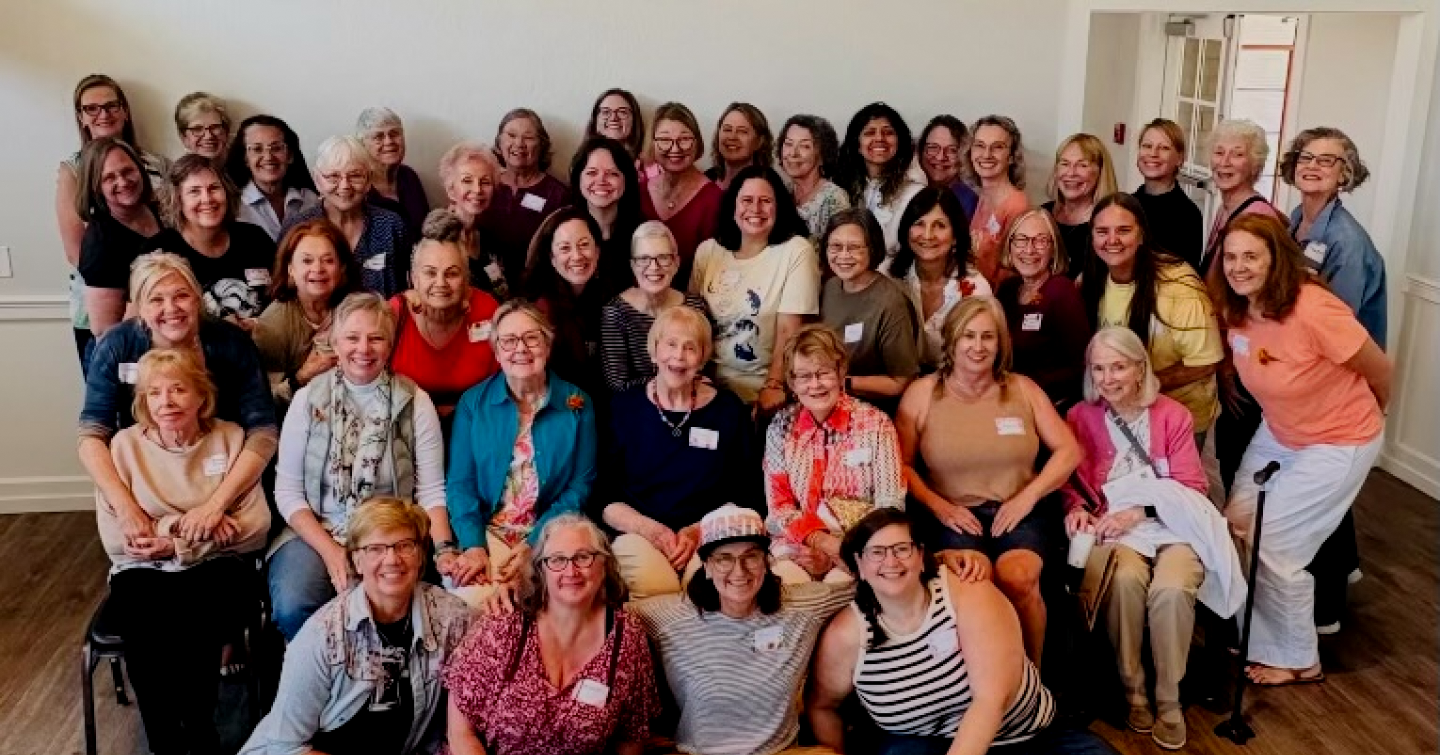 Group of women gathered in a room for a photo, all smiling and standing or sitting closely together.