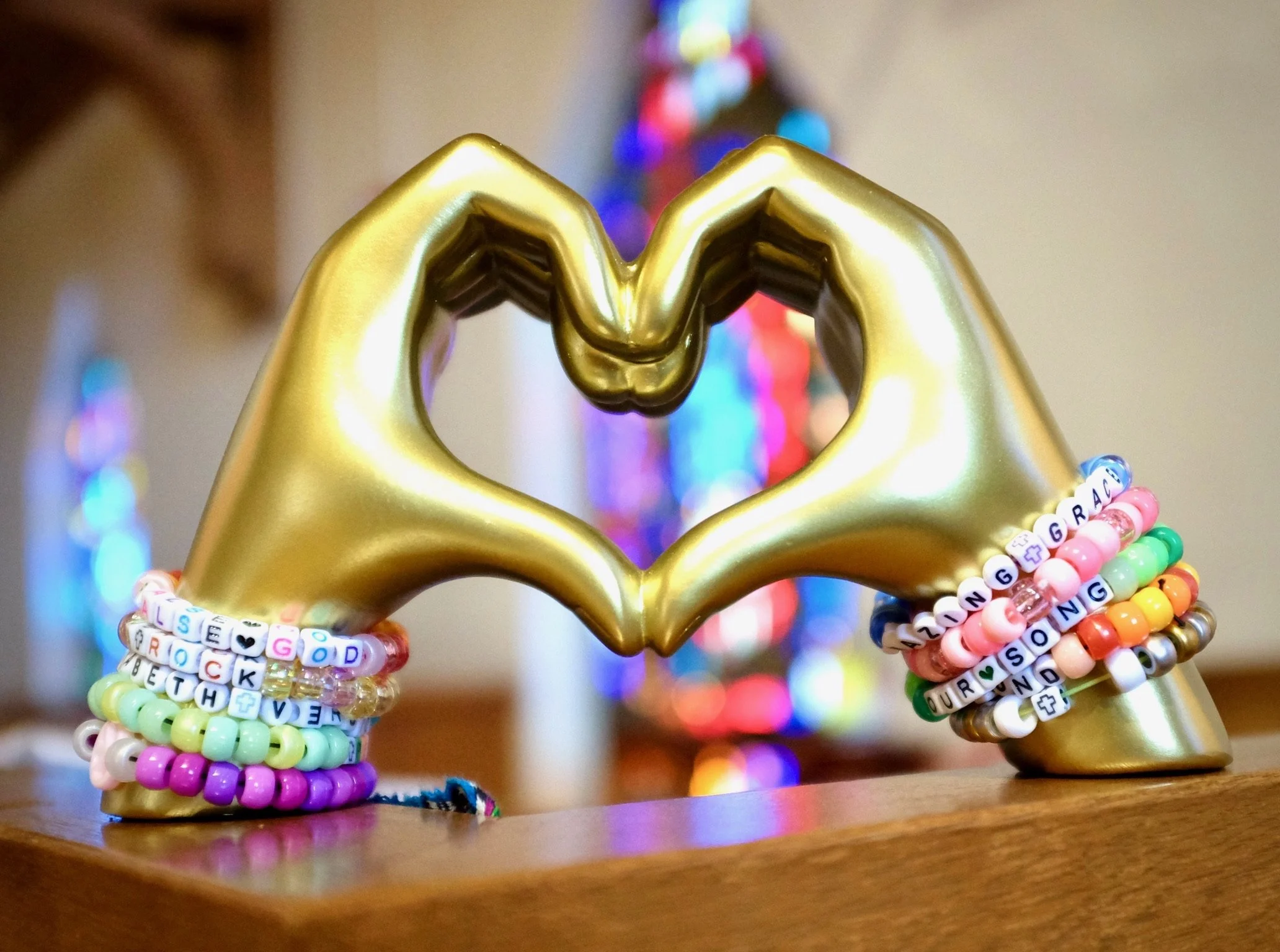 Gold-colored hands shaped into a heart, surrounded by colorful beaded bracelets with words and symbols, positioned on a wooden surface with a blurred colorful Christmas tree in the background.