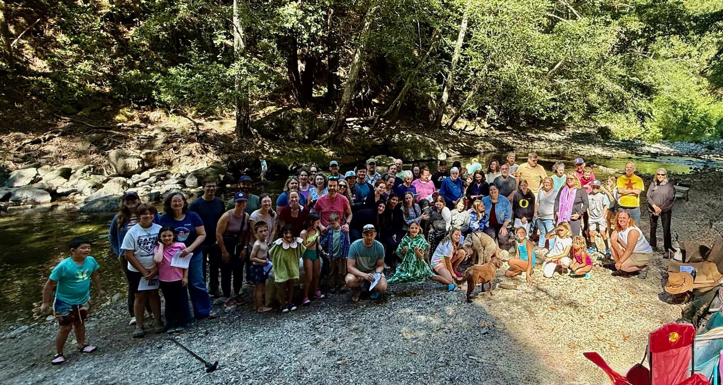 A large group of people gathered outdoors by a river in a wooded area, posing for a group photo. There are children, adults, and elderly individuals, some standing and some sitting on rocks. The scene is shaded by tall trees, and everyone appears relaxed and happy.