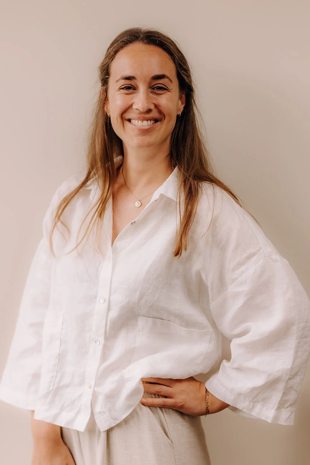 A smiling woman with long brown hair wearing a white linen button-up shirt and light-colored pants, standing against a plain beige background.