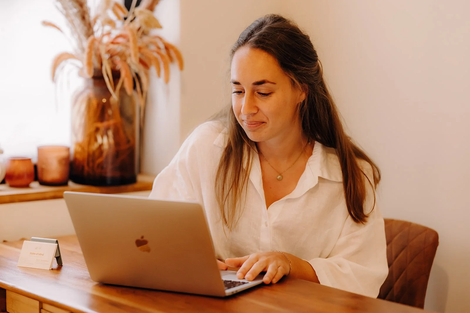 A woman sitting at a wooden table working on a silver MacBook laptop.