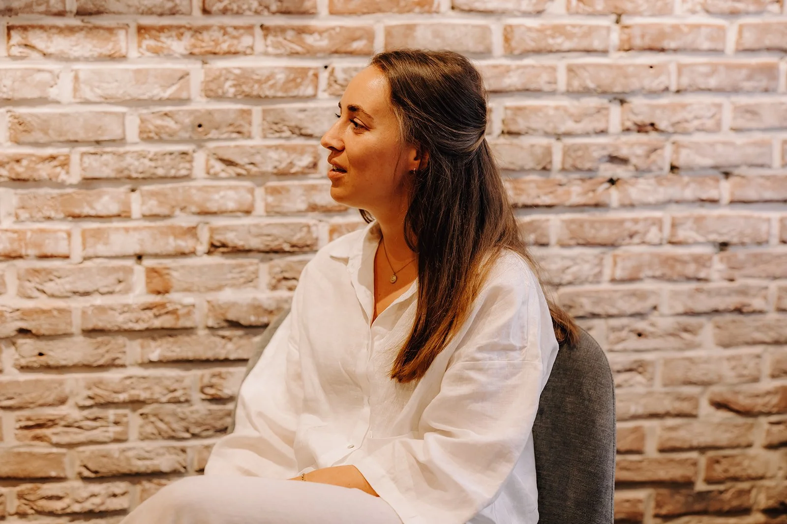 A woman with long brown hair wearing a white shirt sitting on a gray chair against a brick wall.