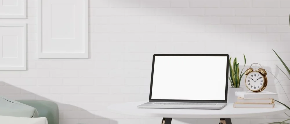 White desk with a laptop, a potted plant, a gold alarm clock, and a small stack of books on it in front of a white brick wall.