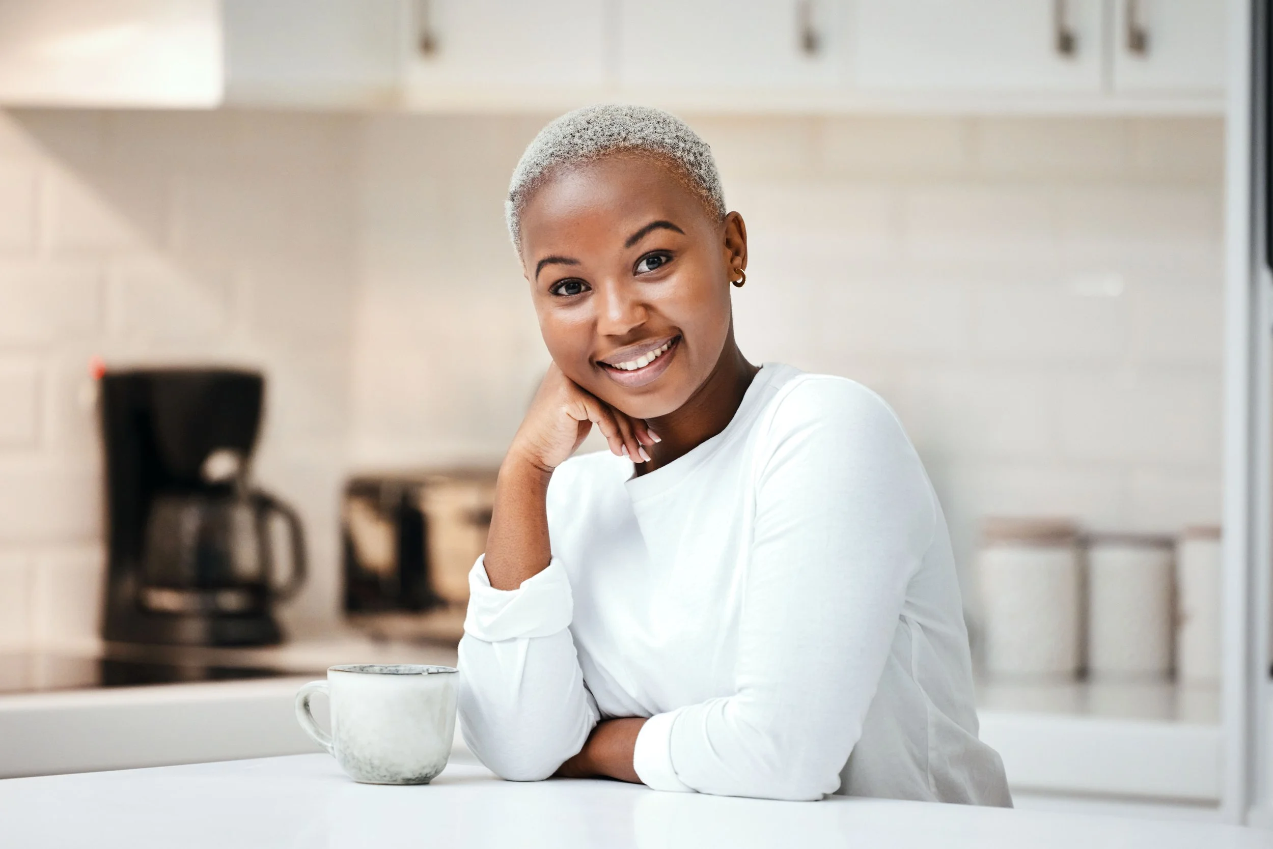 A woman with short blonde hair smiling, sitting at a white kitchen counter with a ceramic mug, in a bright kitchen with white cabinets and a coffee maker in the background.