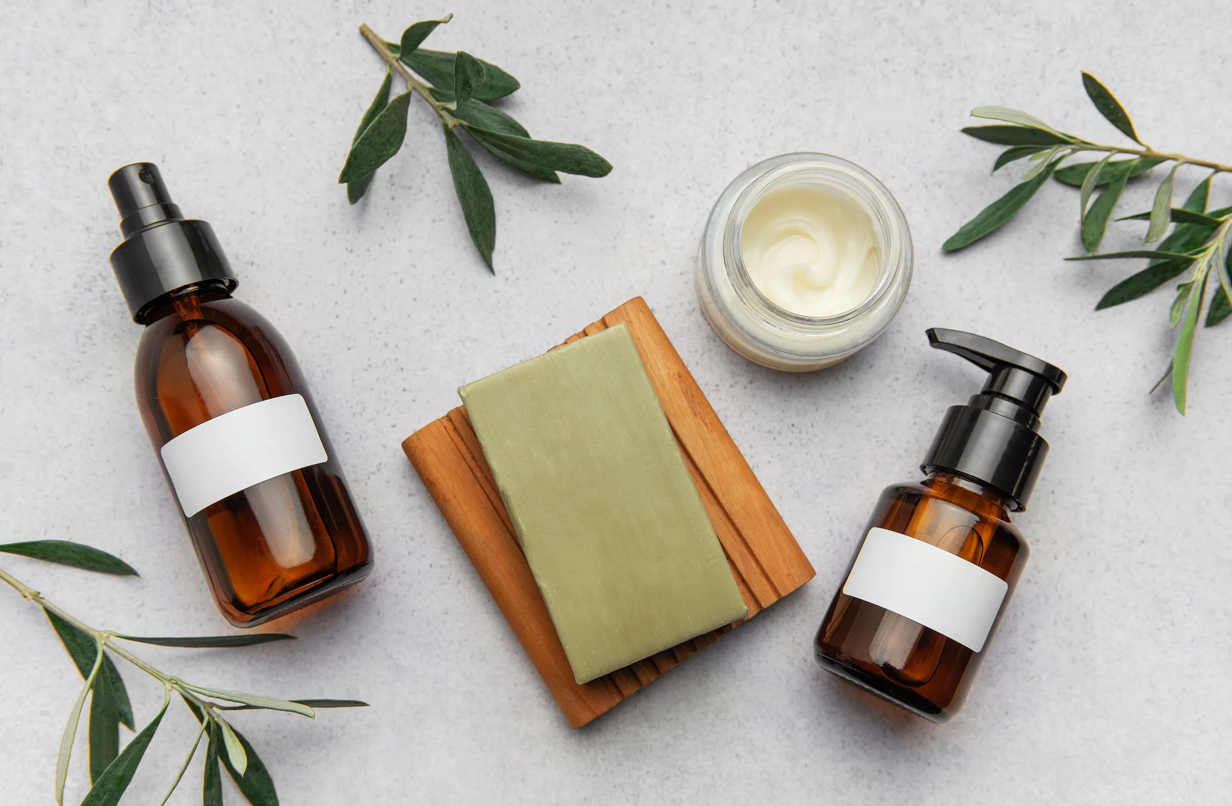 Two amber glass dropper bottles, a green and beige soap bar on a wooden soap dish, a small jar of cream or lotion, and green leafy sprigs on a white surface.