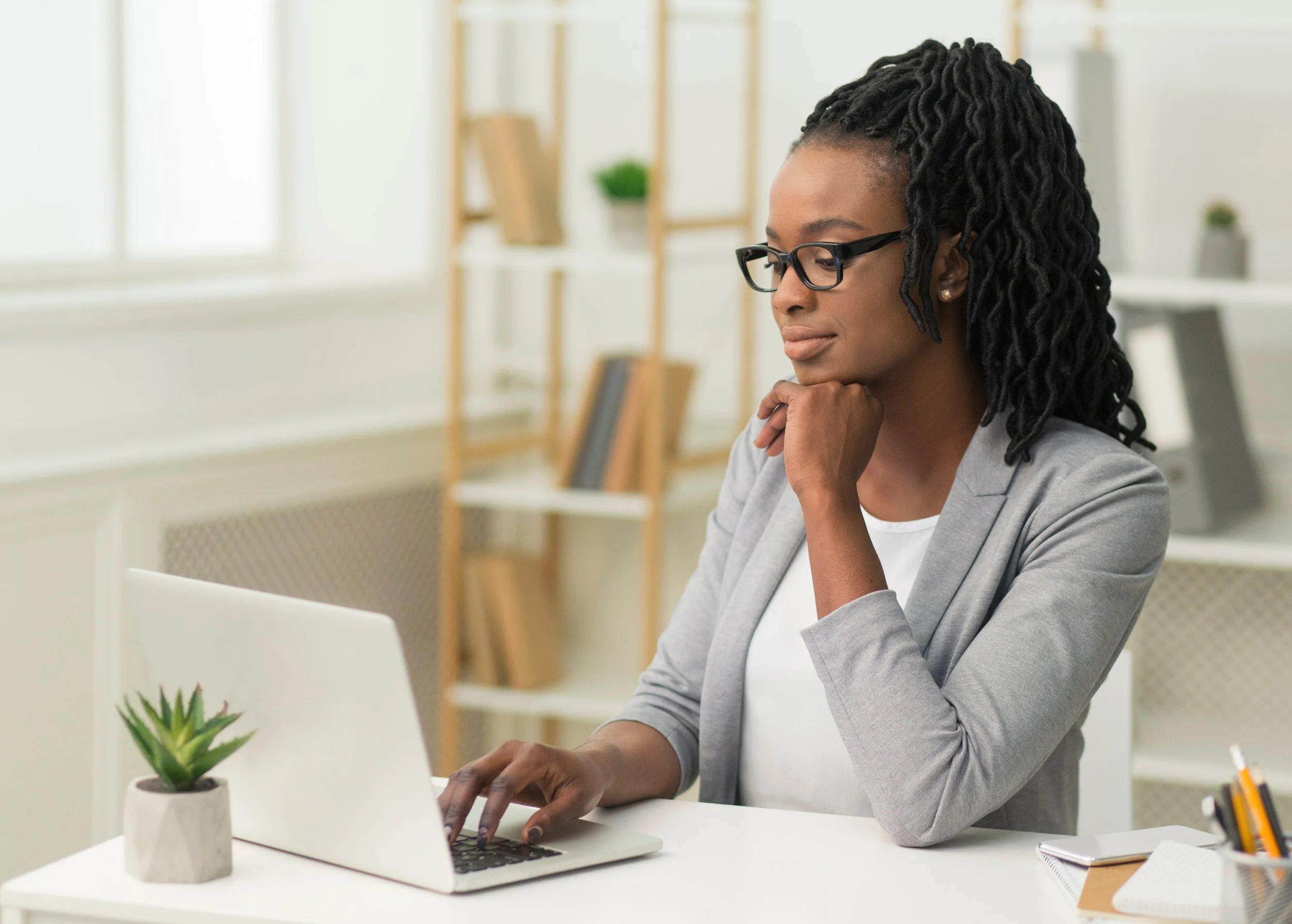 A woman with glasses working on a laptop in an office, with a small potted plant on the desk and shelves with books and decor in the background.