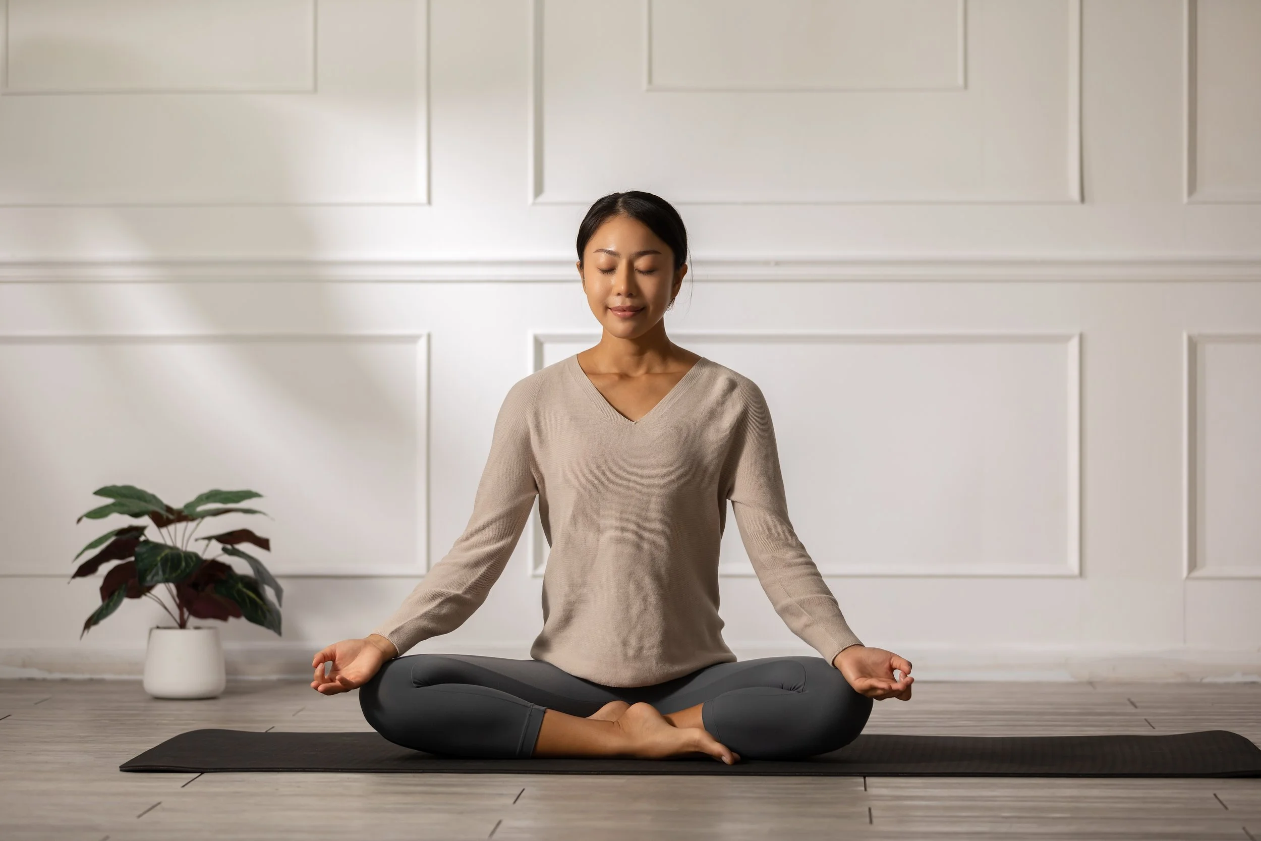 A woman practicing yoga in a sitting cross-legged position on a mat in a bright room with white walls and a potted plant nearby.