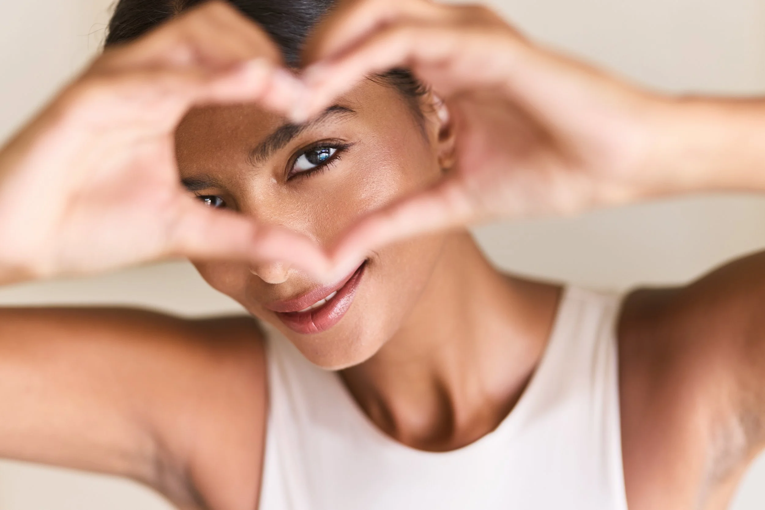 A smiling woman making a heart shape with her hands around her eye, looking through the heart shape.