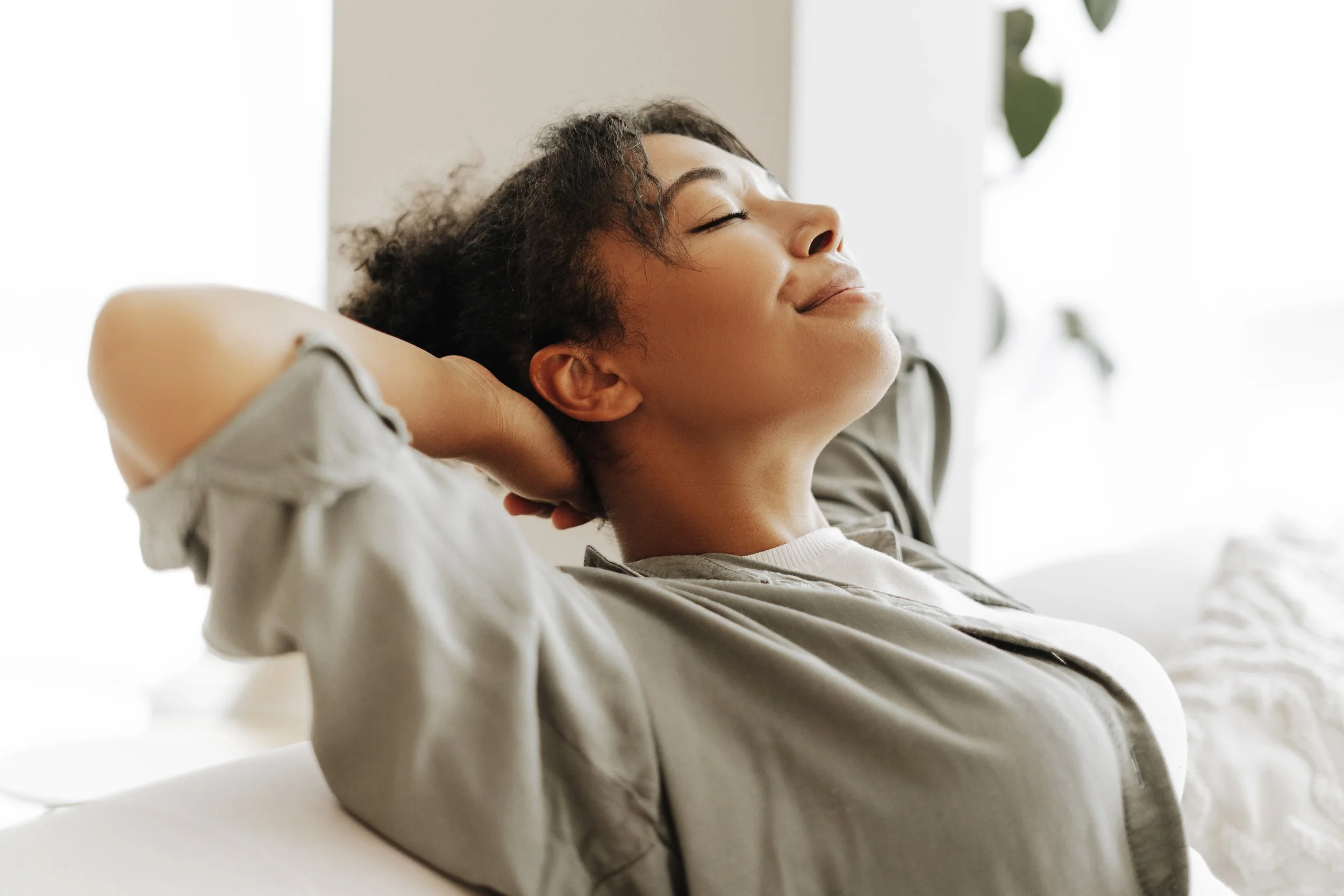 A woman with curly hair, smiling with eyes closed, sitting relaxed on a couch with hands behind her head.