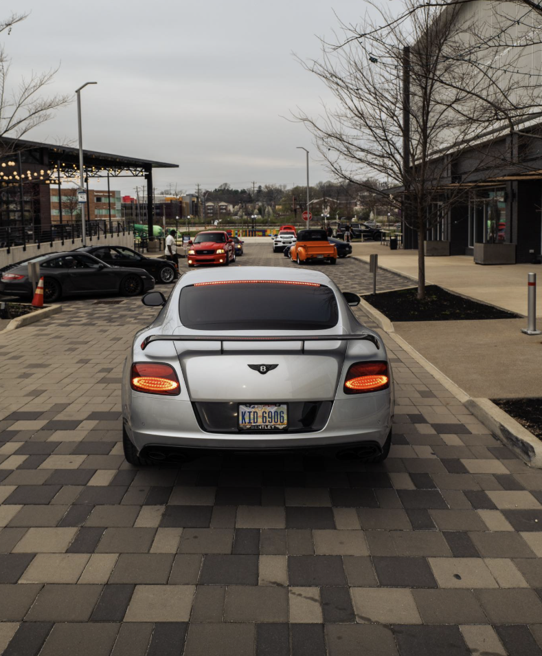 Silver Bentley sports car viewed from the rear in a parking lot with other cars and a shopping area in the background.