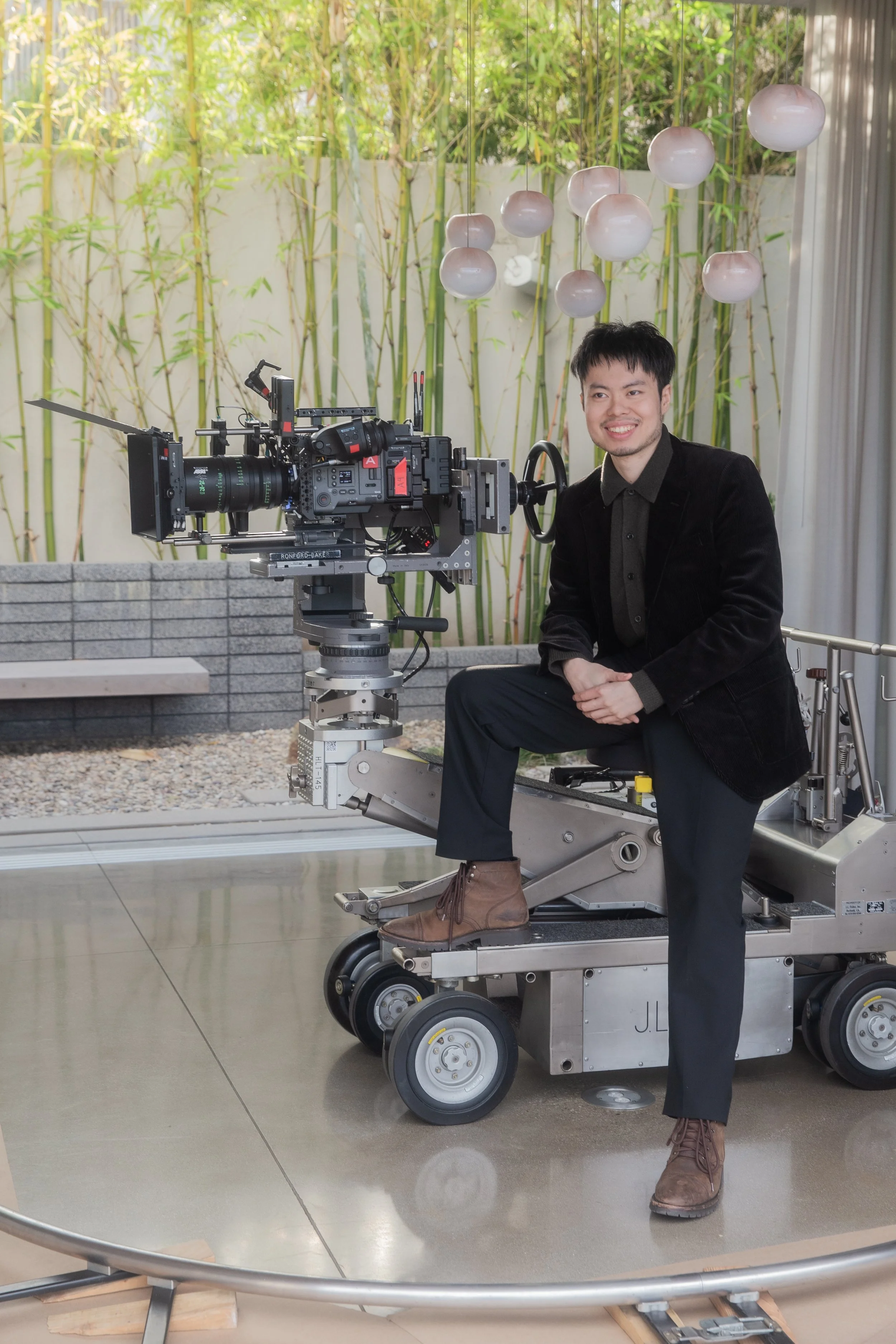 A man sitting on a moving camera dolly with a professional video camera, smiling indoors with a bamboo garden background and decorative hanging lights.