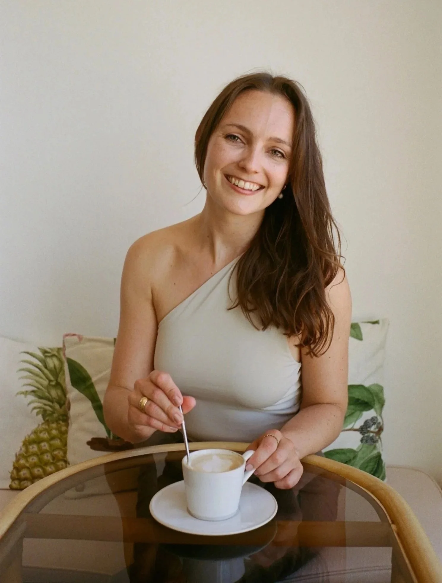 A smiling woman with long brown hair in a white sleeveless top is sitting at a round table, stirring coffee in a white cup. There are tropical fruit illustrations on a cushion behind her, including pineapples and leaves. Rebecca Steinke is pictured.