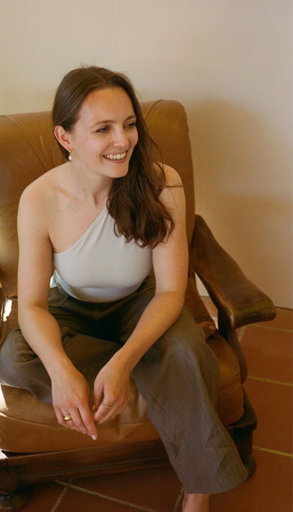 A woman with long brown hair, wearing a white sleeveless top and brown plaid pants, sitting on a vintage wooden armchair with a smile. Rebecca Steinke is pictured.