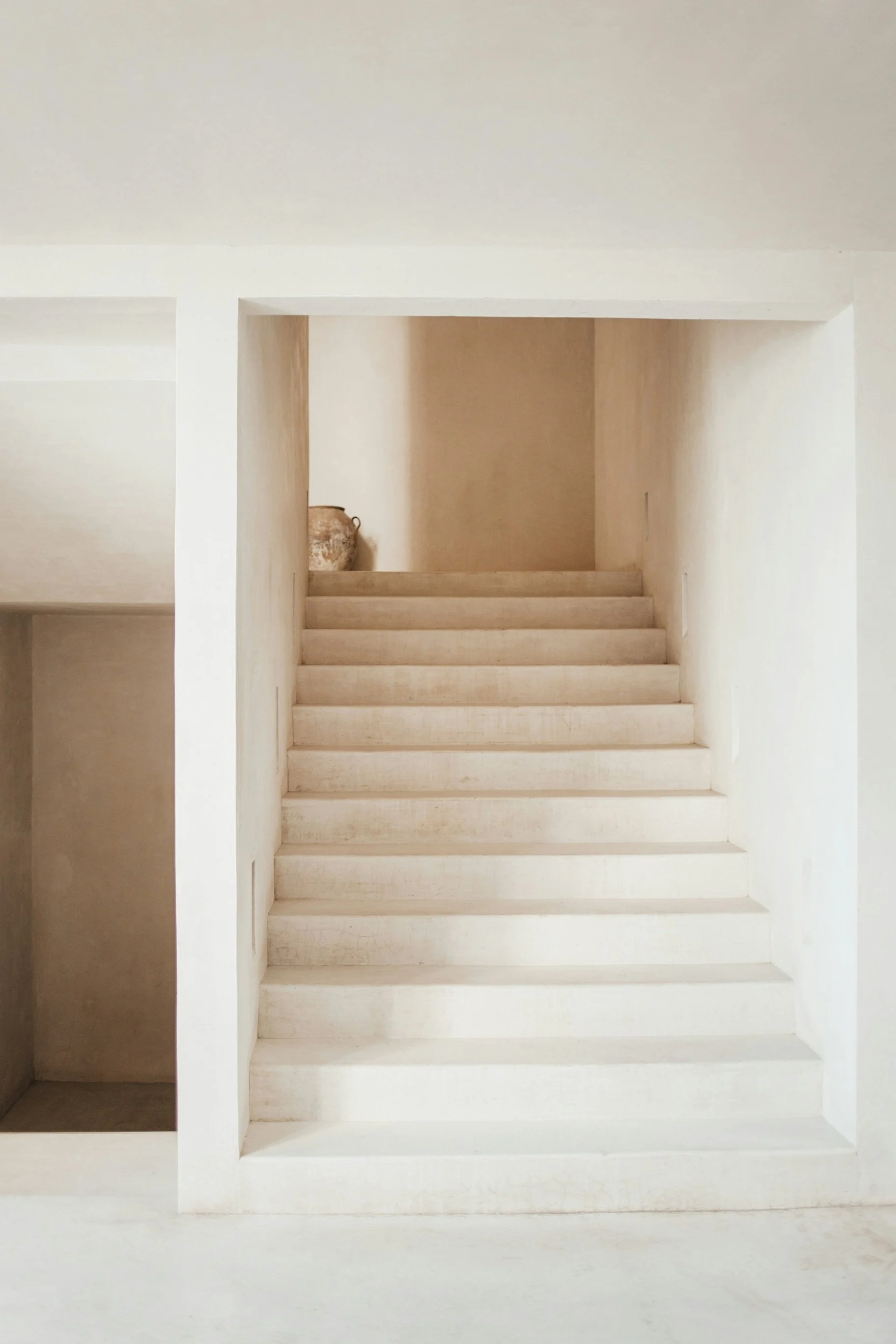 Minimalist staircase with light-colored steps and walls, featuring a large decorative urn on the landing to the left.