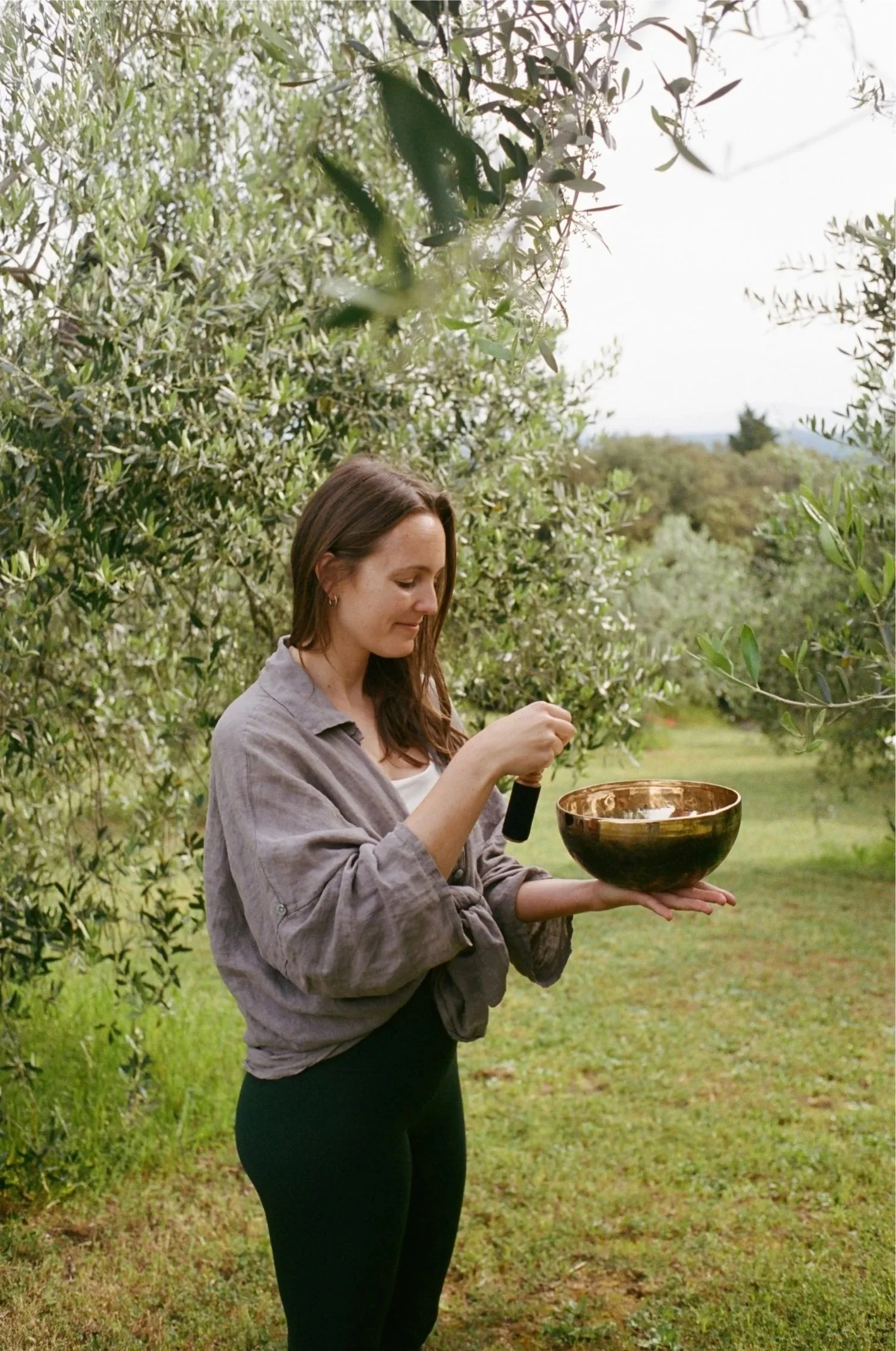 A woman with brown hair wearing a gray linen shirt and darl green pants holding a singing bowl outdoors in a green garden with olive trees and hills in the background. Rebecca Steinke is pictured.