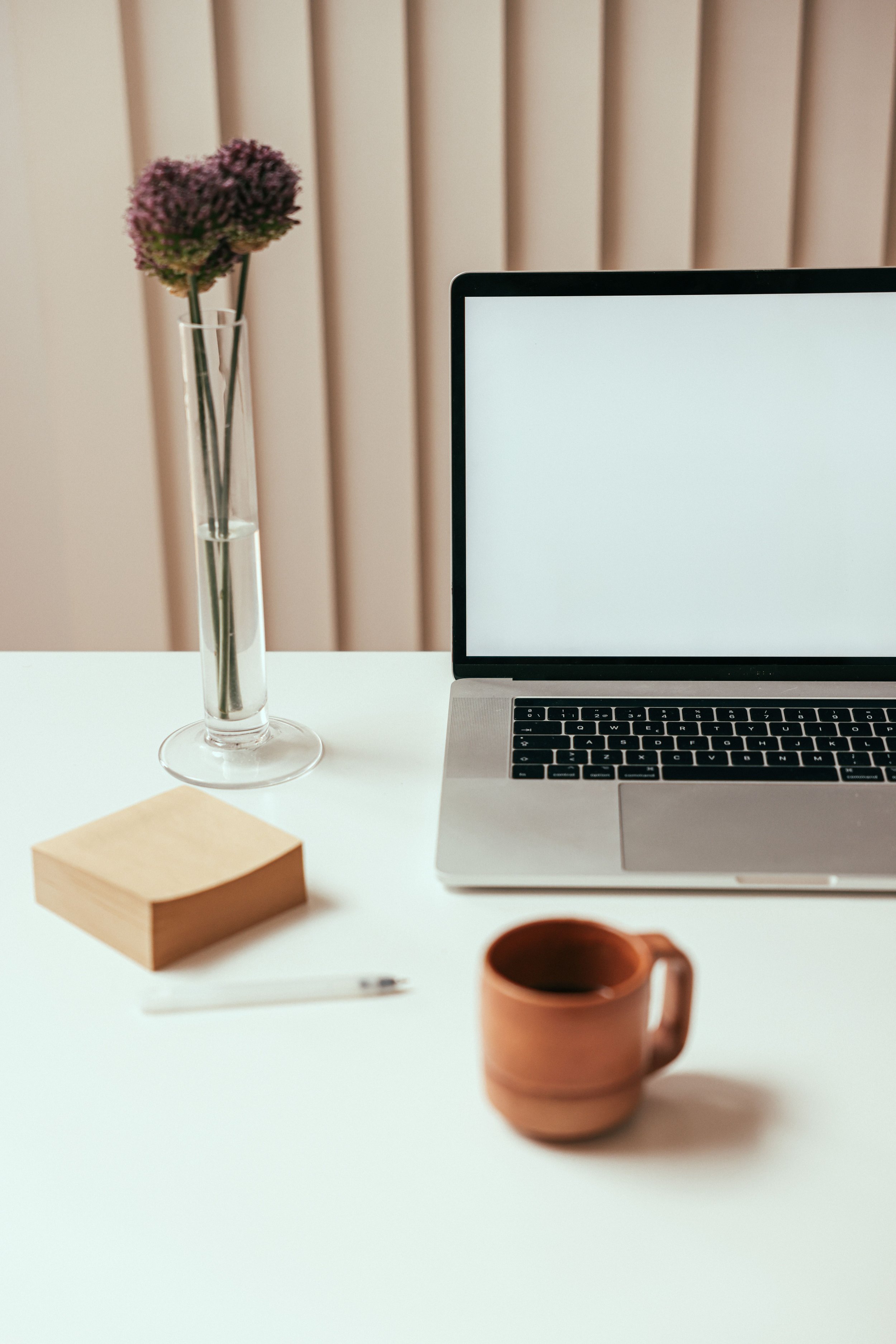 A workspace with a silver laptop, a tall vase of purple flowers, a small wooden box, a white pen, a brown mug, and a sliding door behind.