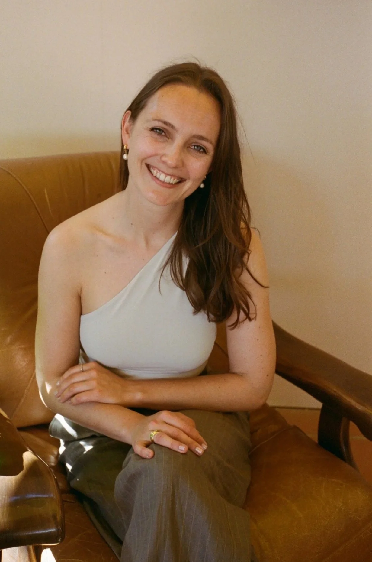 A smiling woman with long reddish-brown hair, wearing pearl earrings and a white one-shoulder top, sitting on a brown leather bench. Rebecca Steinke is pictured.