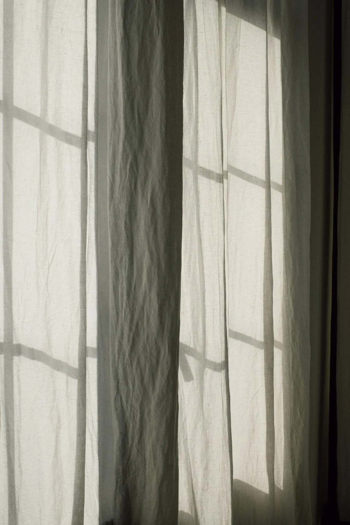 Sunlight filters through semi-transparent white and beige curtains, casting shadows from a window frame.