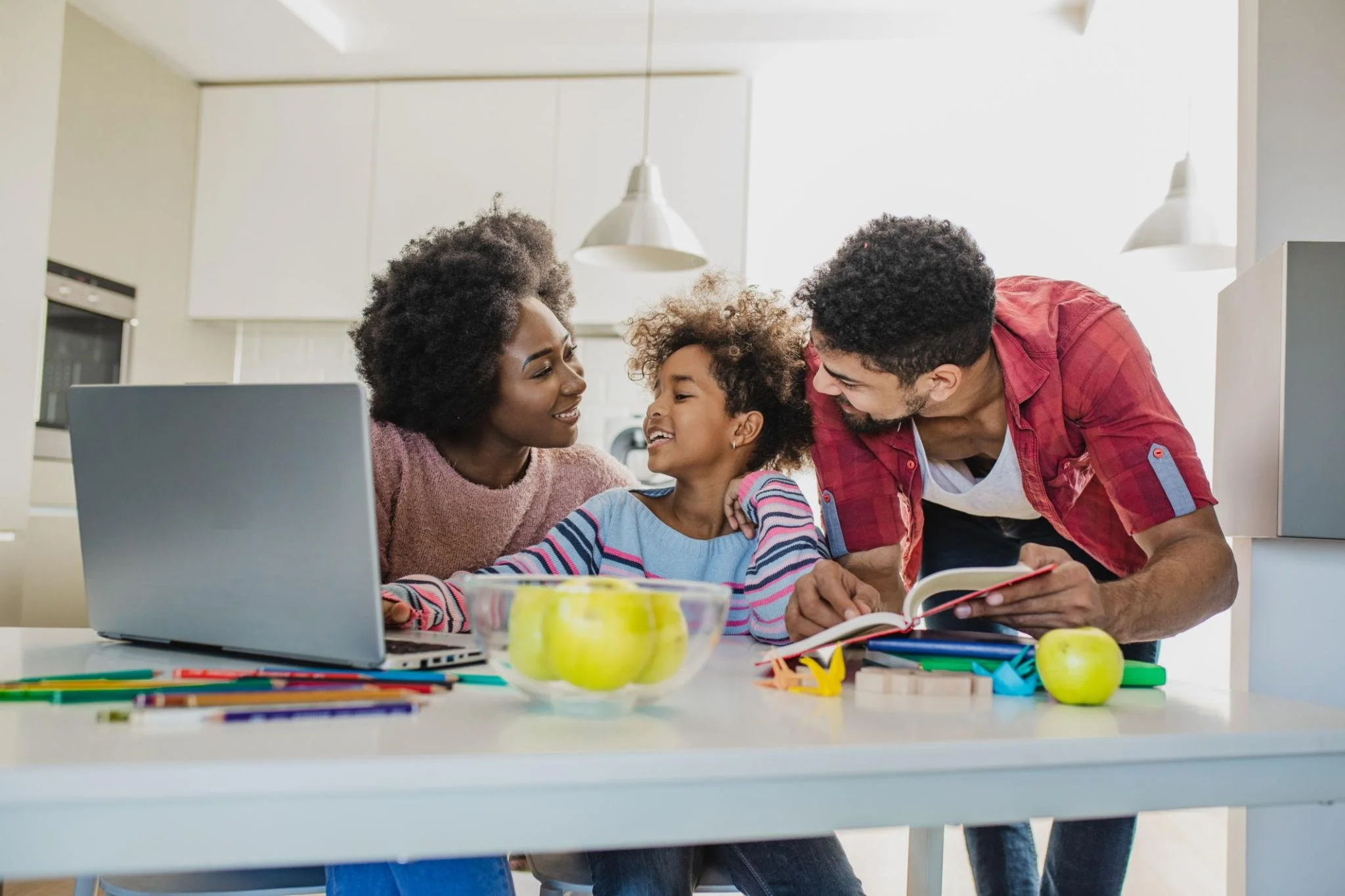Family of three gathered around a table in a bright kitchen, smiling. The mother and father are leaning in towards their young daughter, sharing a happy moment. The table has school supplies and a laptop, suggesting they are doing homework or studying together.