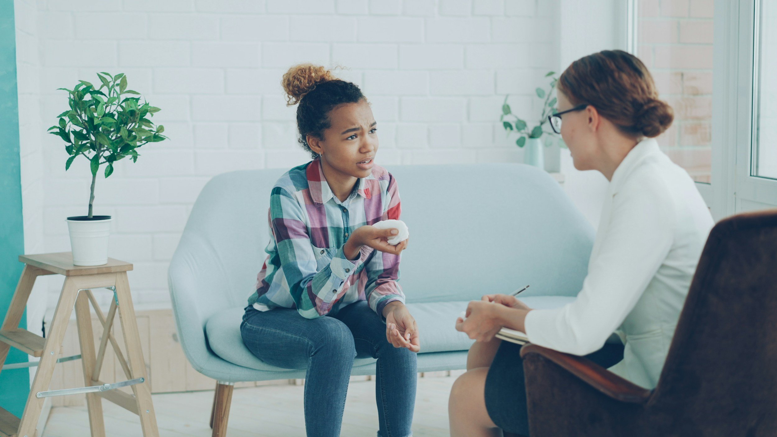 Young woman with curly hair and plaid shirt sitting on a couch, talking seriously with a female therapist in a white coat, during a counseling session in a bright room with white brick walls and potted plants.