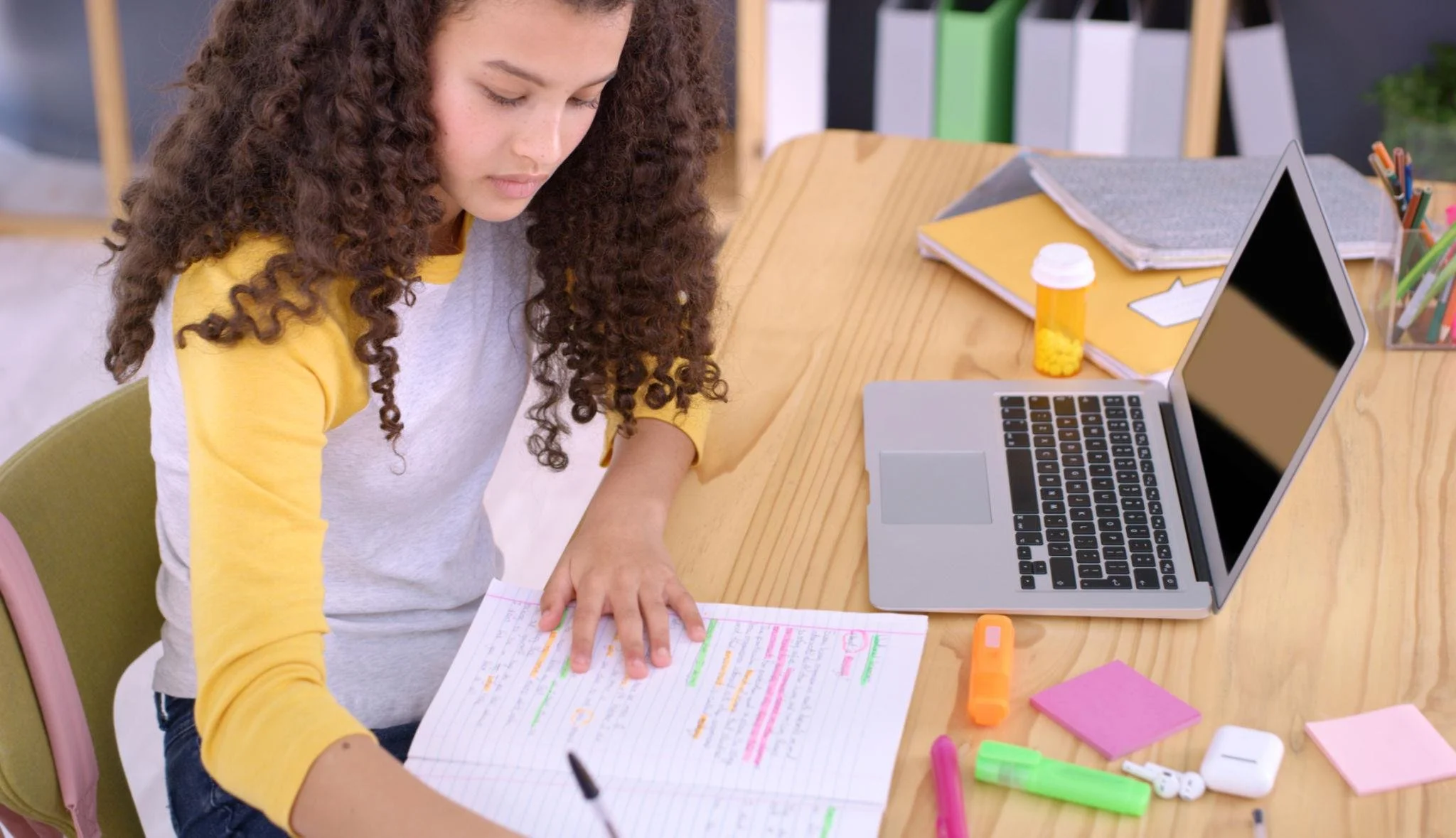 A young woman with curly hair sitting at a desk, studying with colorful highlighters and sticky notes, with a laptop and notebooks nearby.